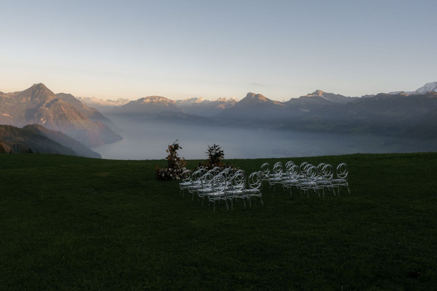 White chairs arranged on a mountain meadow at dusk with misty Alps and a glowing horizon in the background.