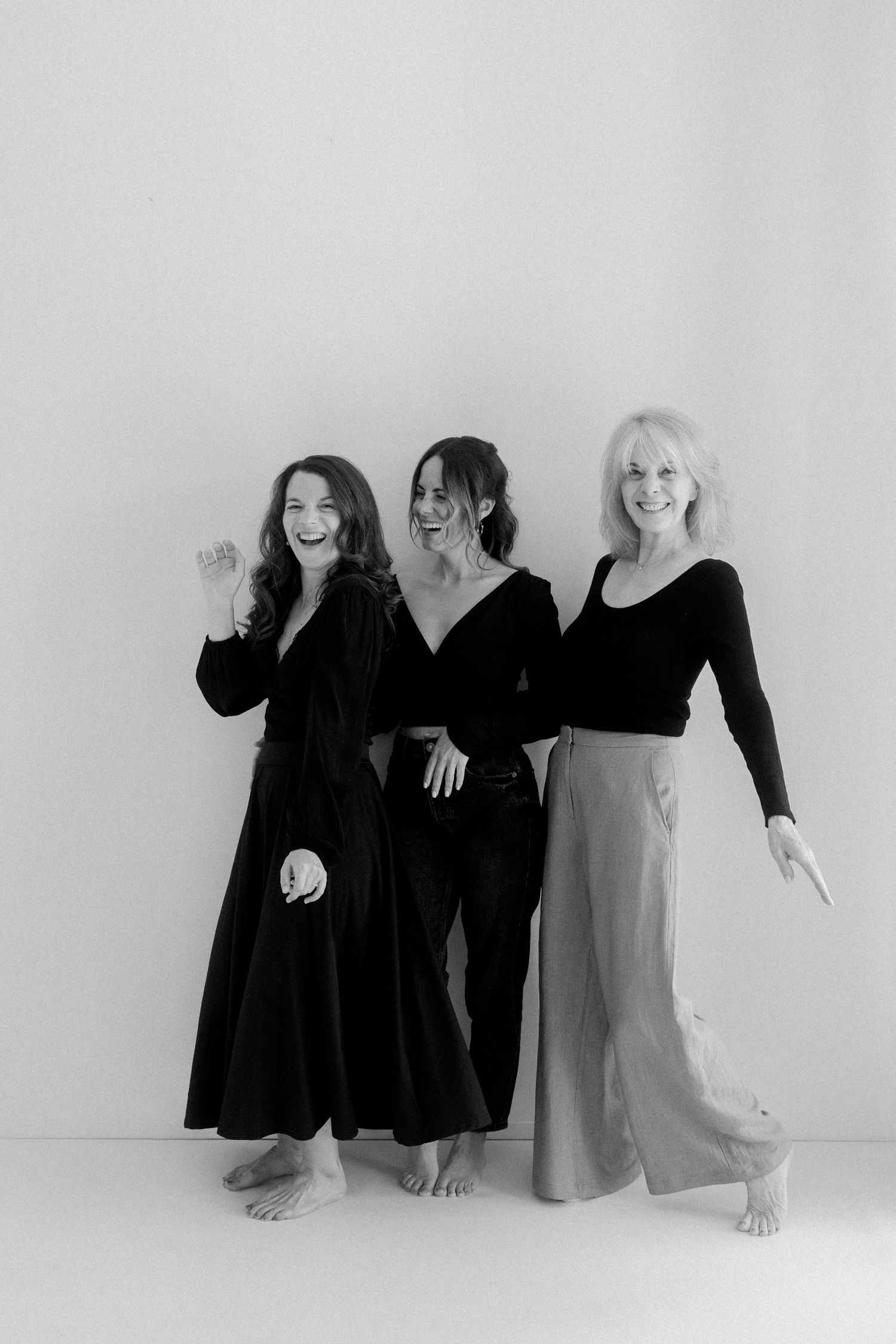Three women in black and neutral clothing laughing and posing together in a minimalist black and white portrait.