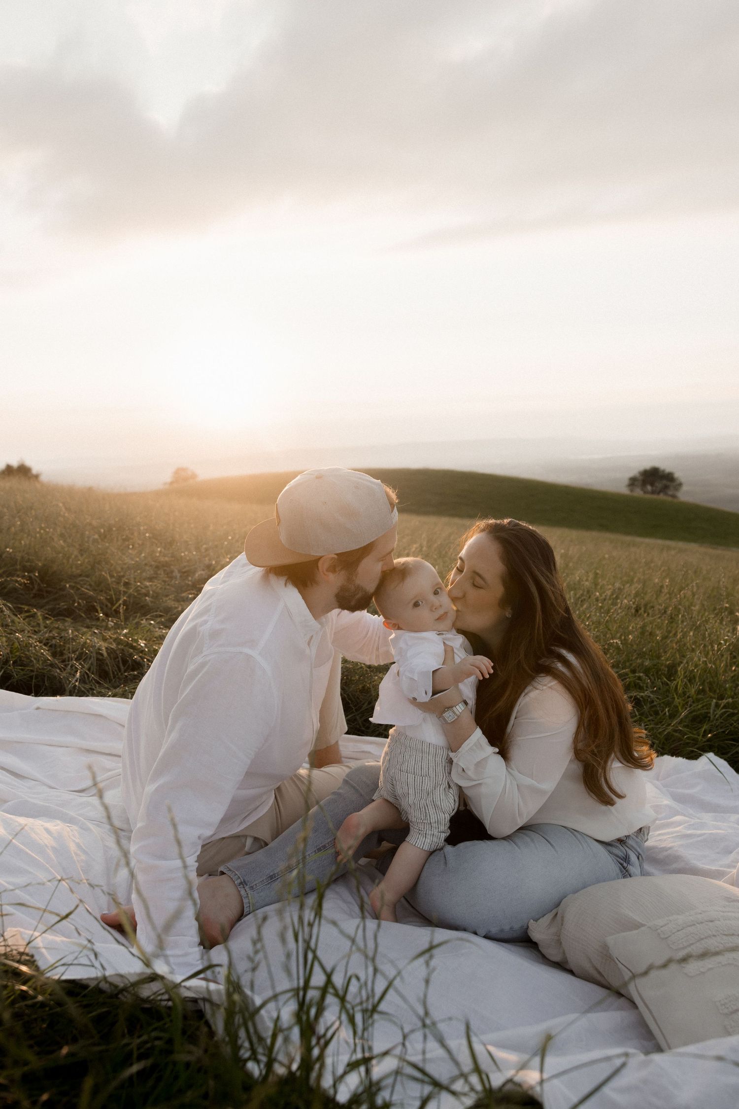 Family lying together on white blanket in field during golden sunset.