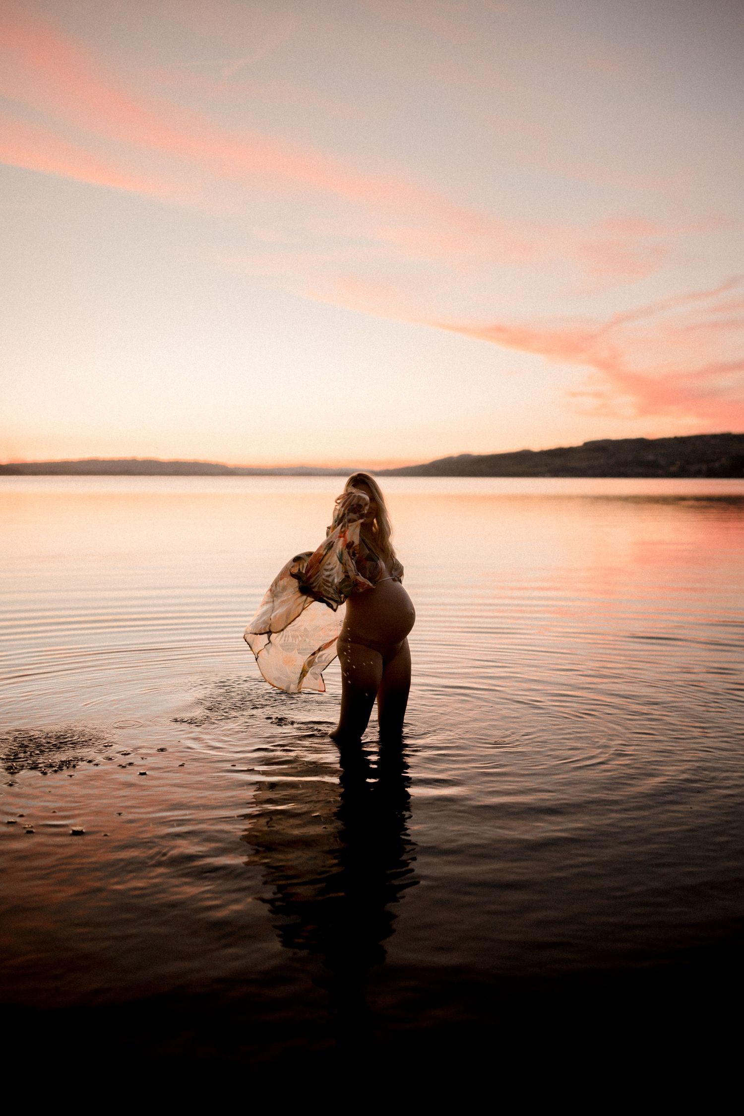 Person standing in calm lake water during vibrant sunset with mountains in background.