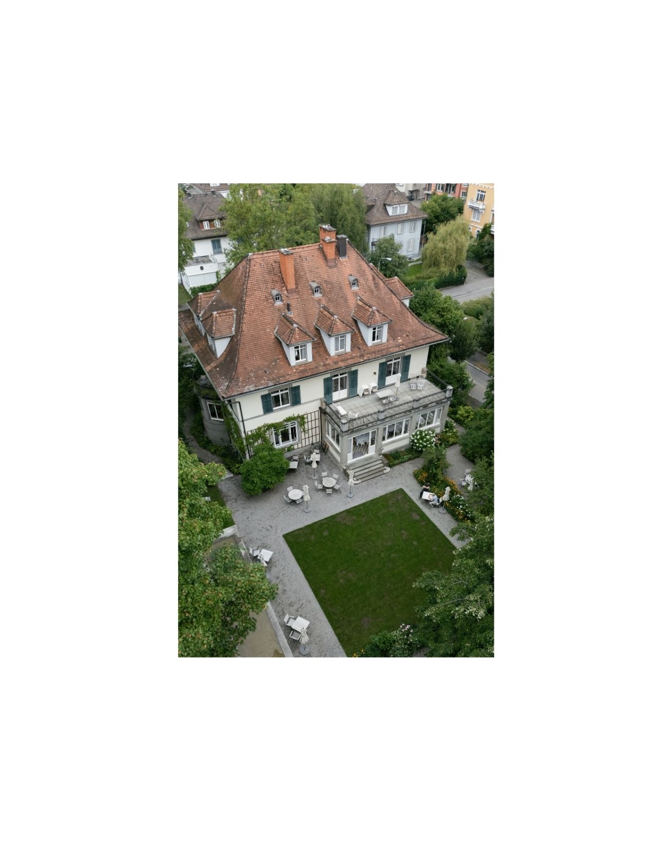 Aerial view of a charming European villa with red tile roof, green shutters, manicured lawn, and surrounding mature trees.