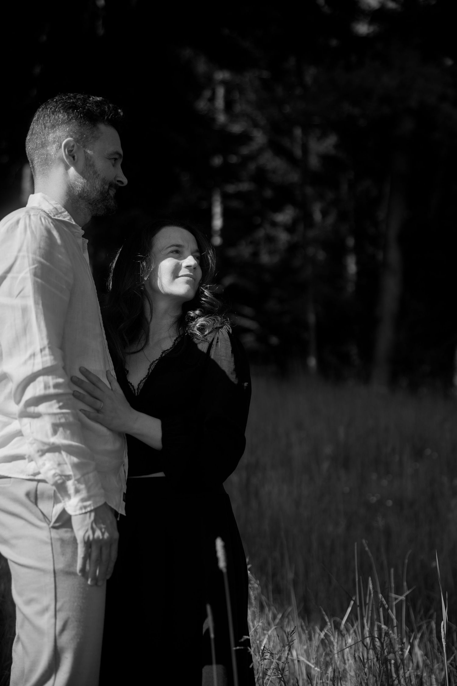 Black and white romantic engagement photo series showing couple embracing in a sunlit meadow at sunset.