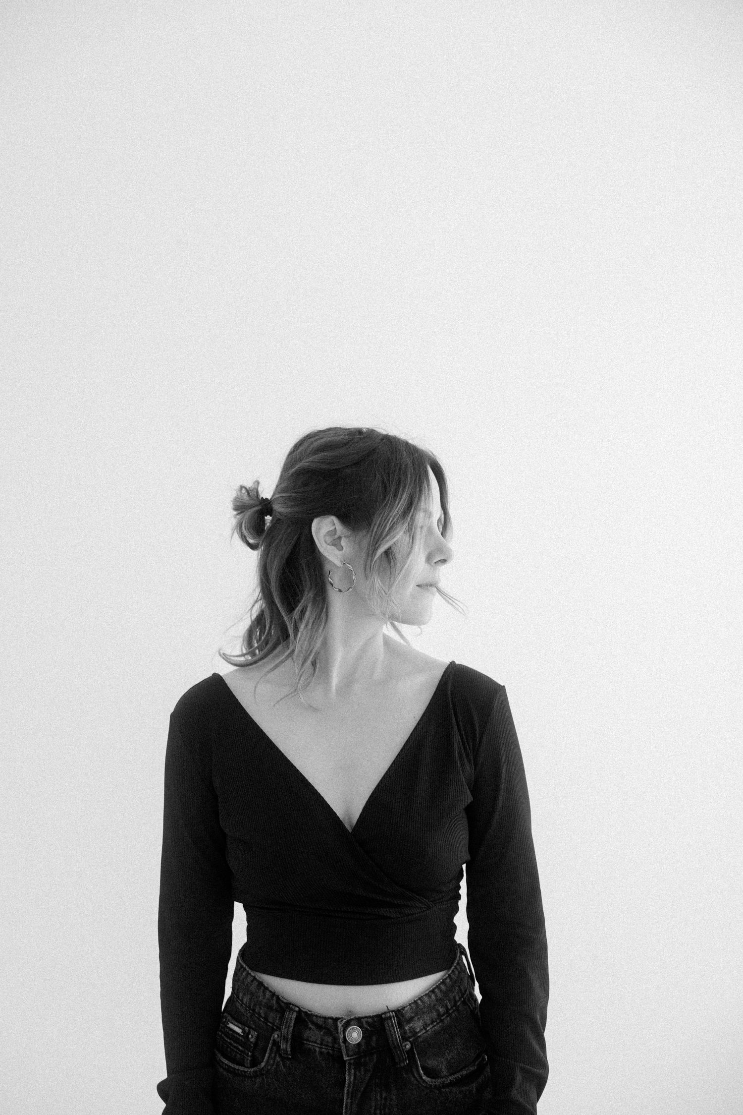 Black and white portrait of person in v-neck crop top and jeans looking to the side against white wall.