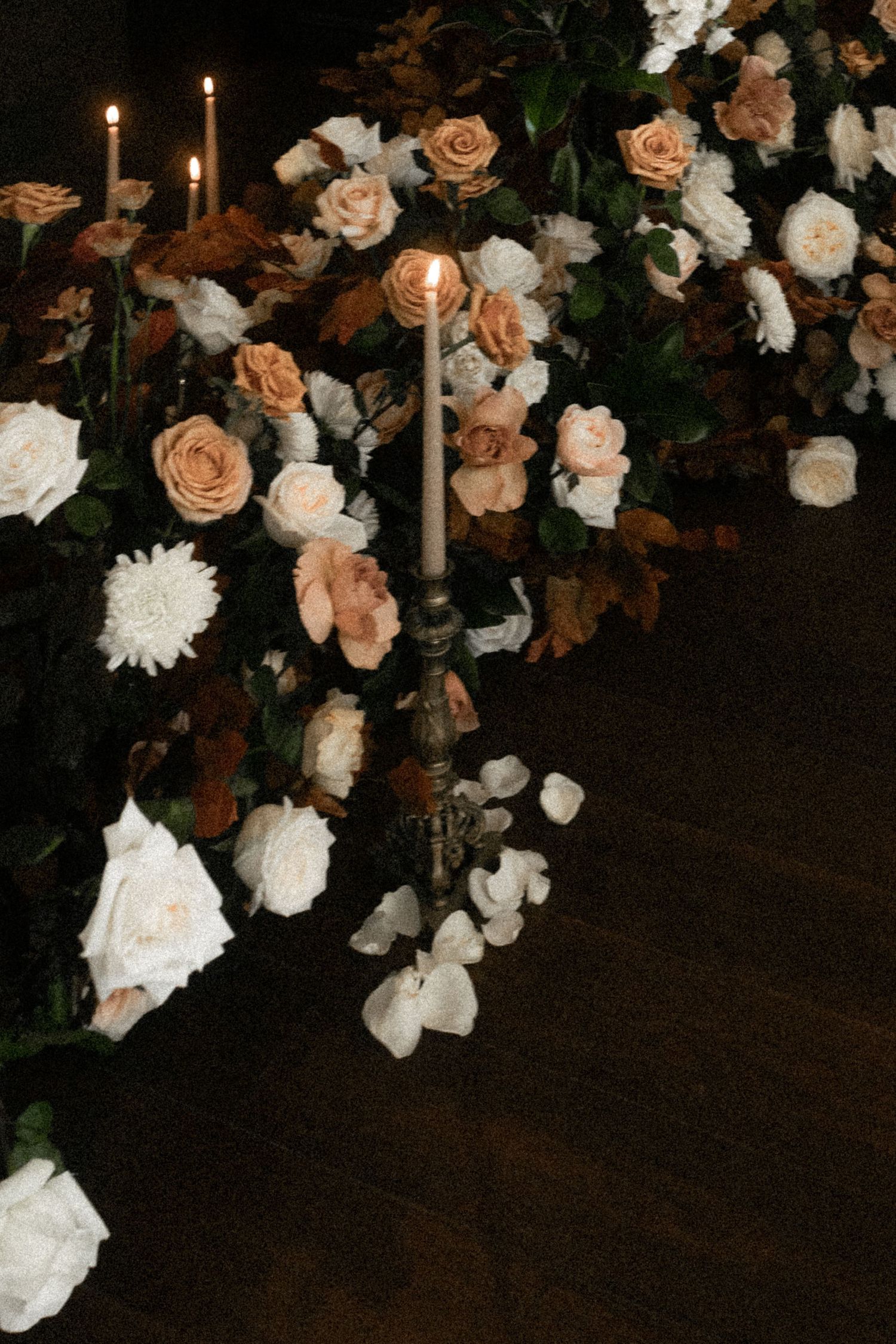 Elegant candlelit floral arrangement with peach and white roses surrounding ornate candelabra in dark romantic setting.