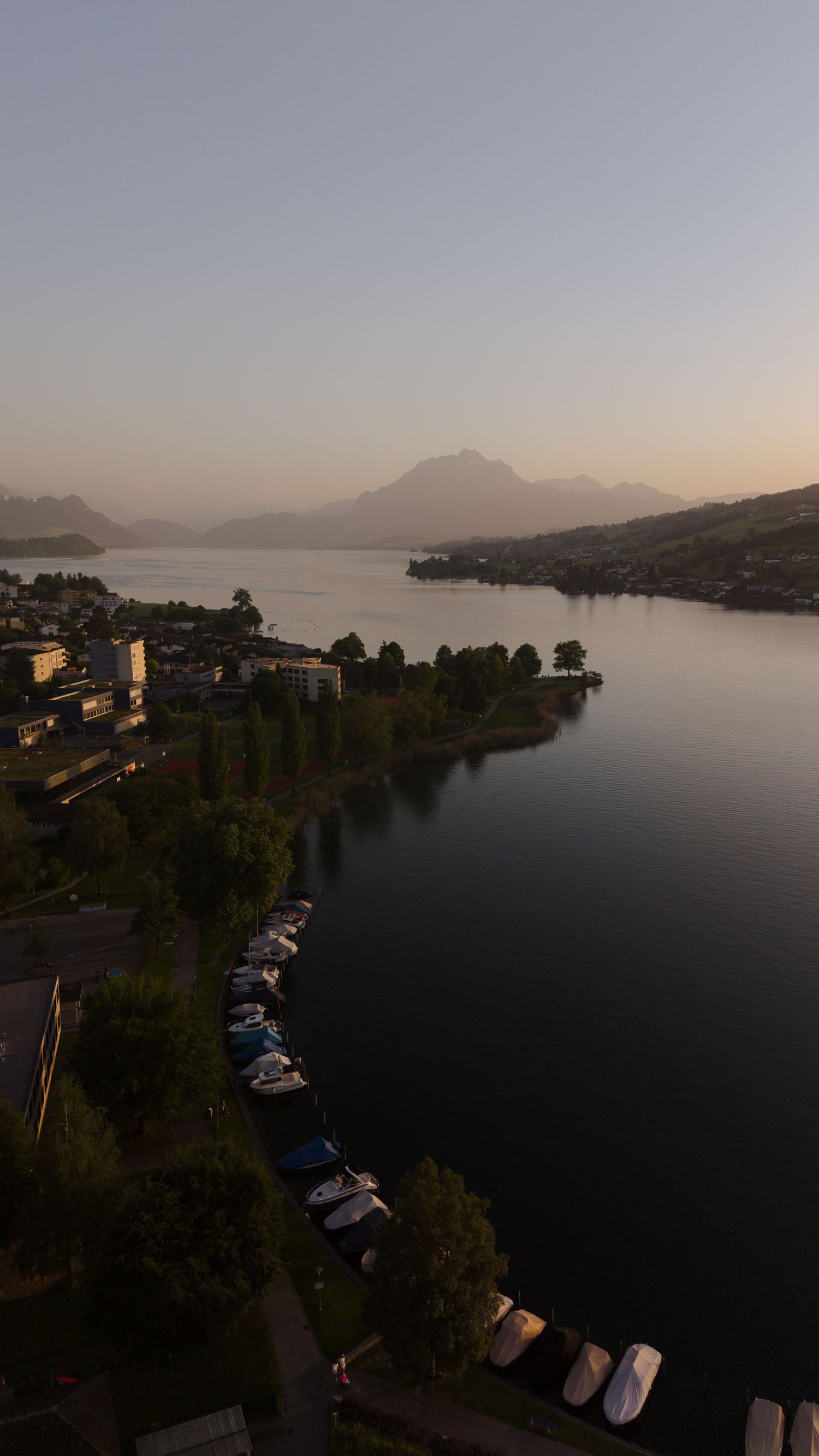 Aerial sunset view of a serene alpine lake with moored boats, lush trees, mountain silhouettes, and a small lakeside town.