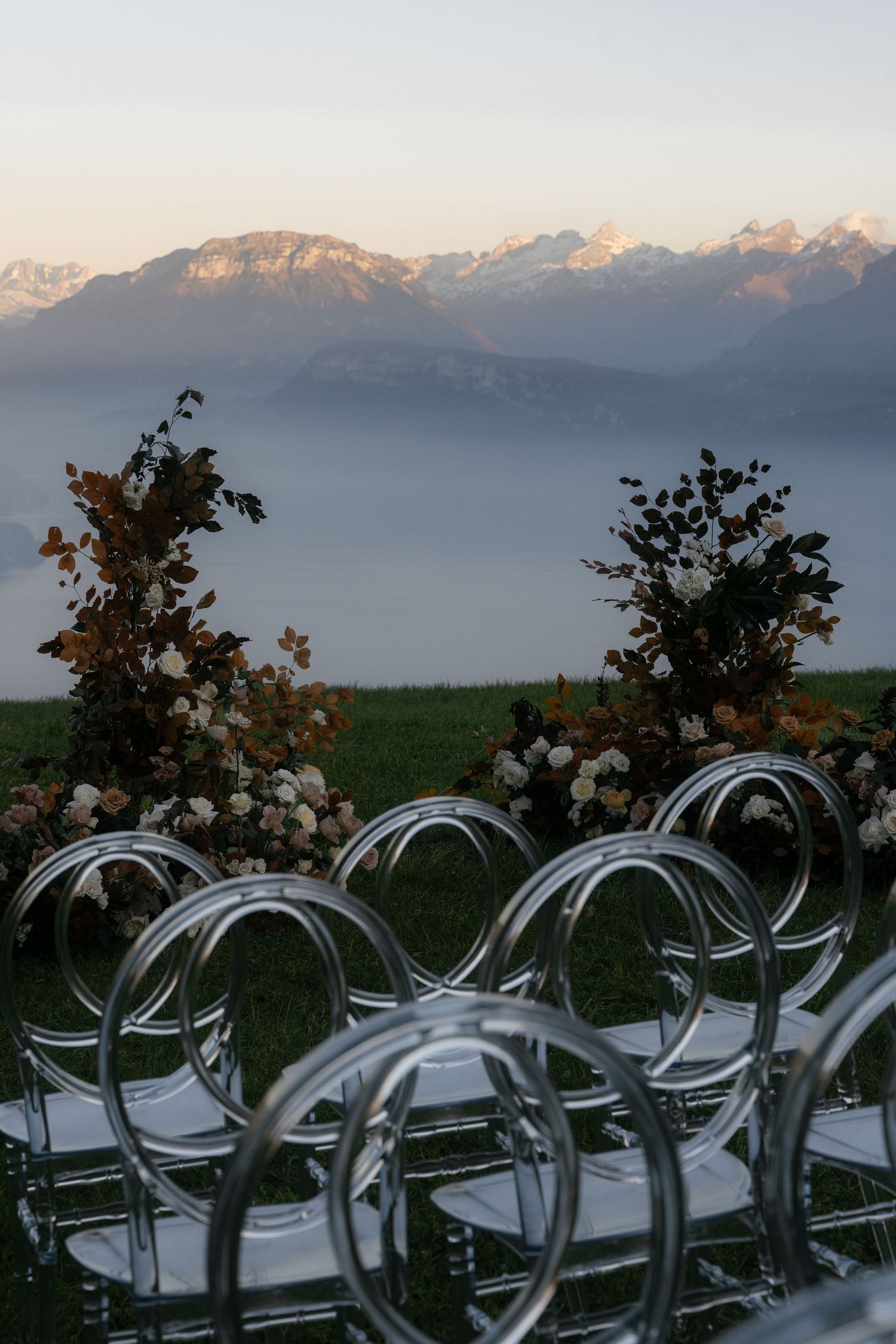 Outdoor wedding ceremony setup with acrylic chairs and floral arrangements against a stunning alpine mountain backdrop at dusk Villa Honegg