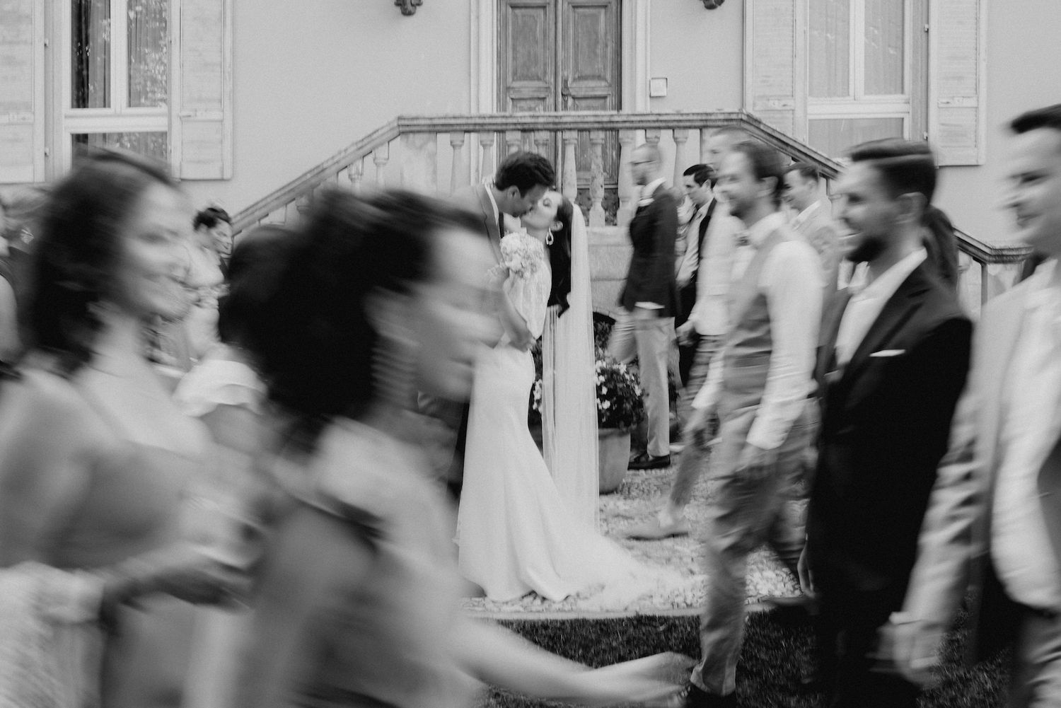 Black and white wedding photo of bride and groom kissing while blurred guests walk past outdoors.