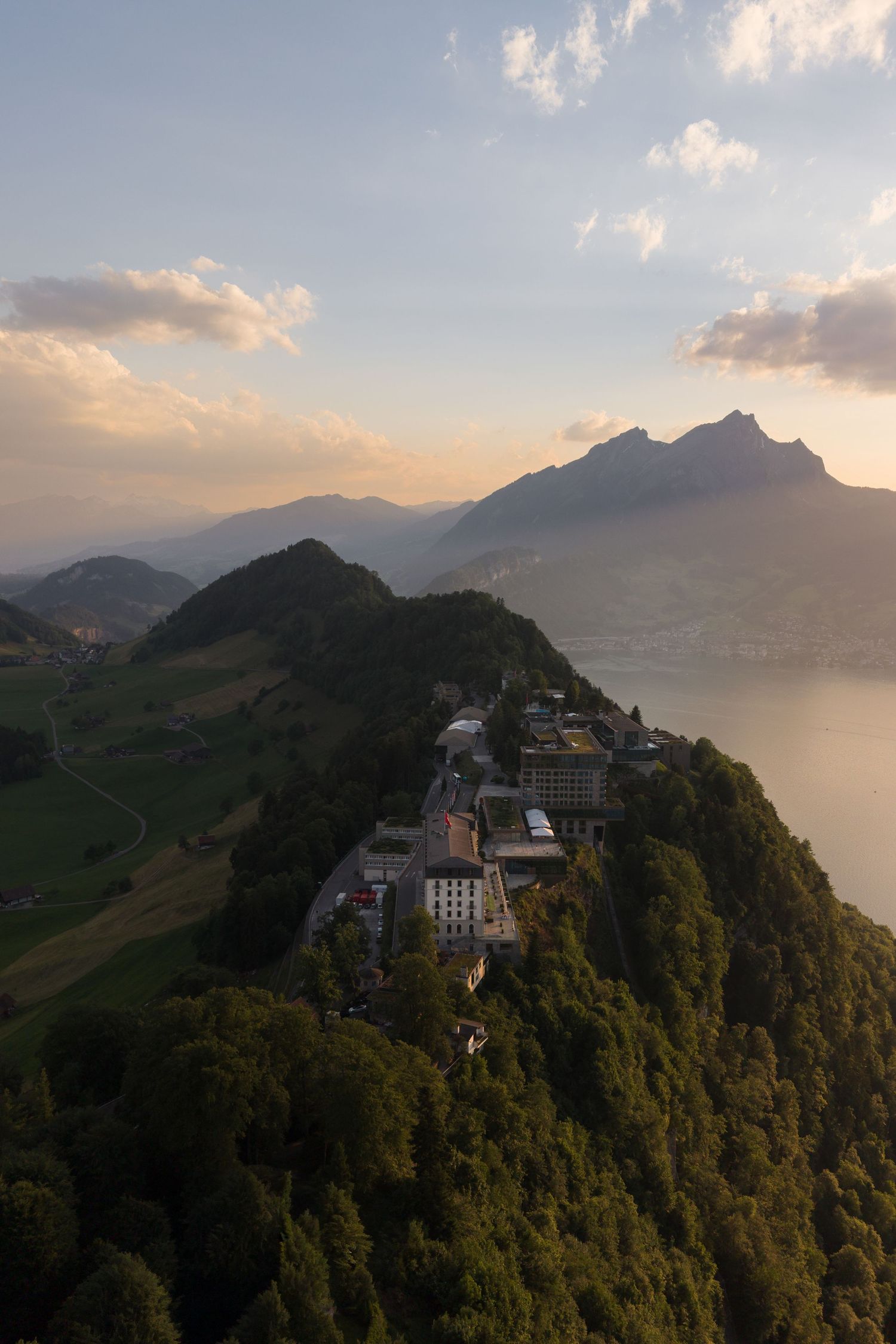 Aerial sunset view of a mountain village perched on a forested ridge overlooking a Swiss alpine lake.