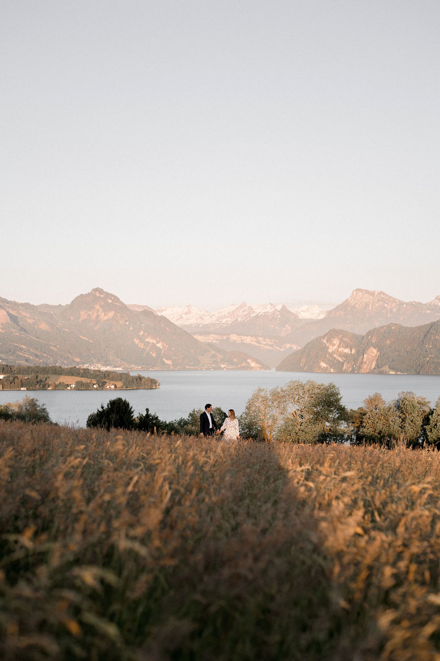 Two people stand in golden grass overlooking a scenic mountain lake at sunset with snow-capped peaks in the distance.