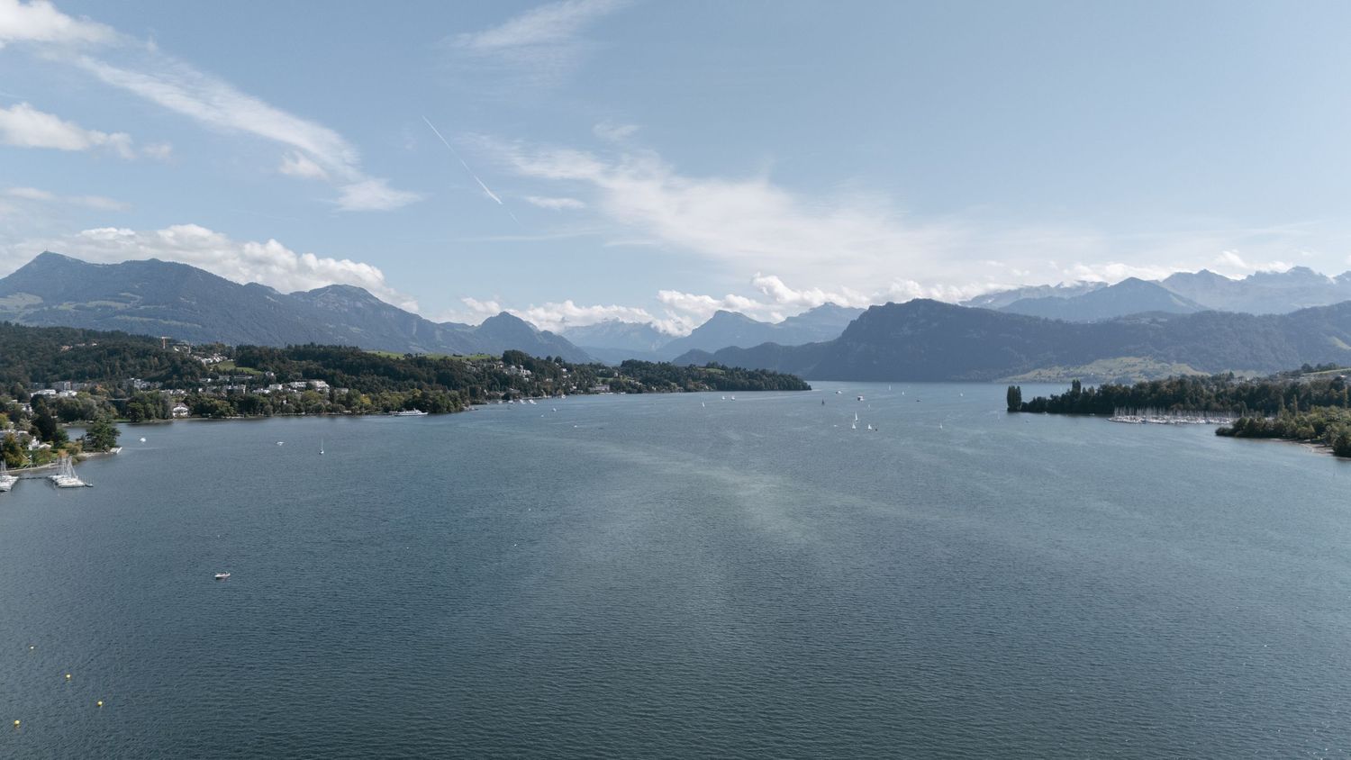 Aerial view of a serene alpine lake surrounded by mountains and lush greenery under a partly cloudy blue sky.