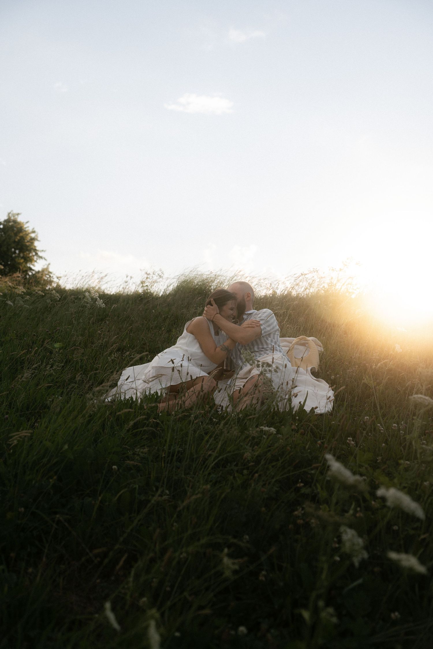 A romantic couple in white wedding attire embraces in a golden sunlit meadow at sunset.