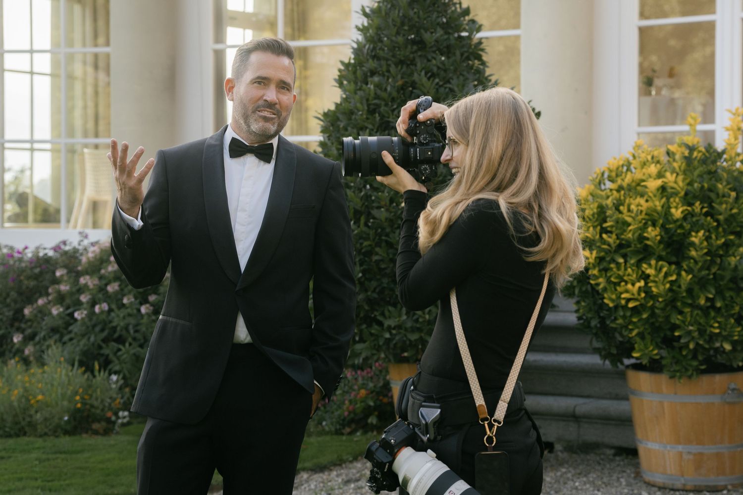 A photographer captures a man in a tuxedo outdoors at an elegant venue with lush greenery.