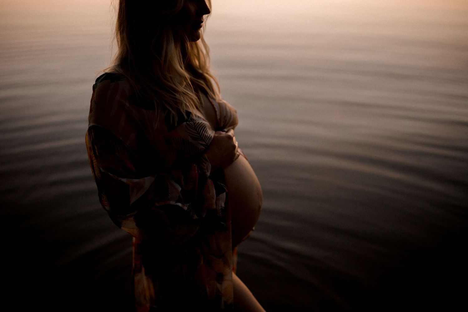 Silhouette of pregnant figure standing in water during sunset.