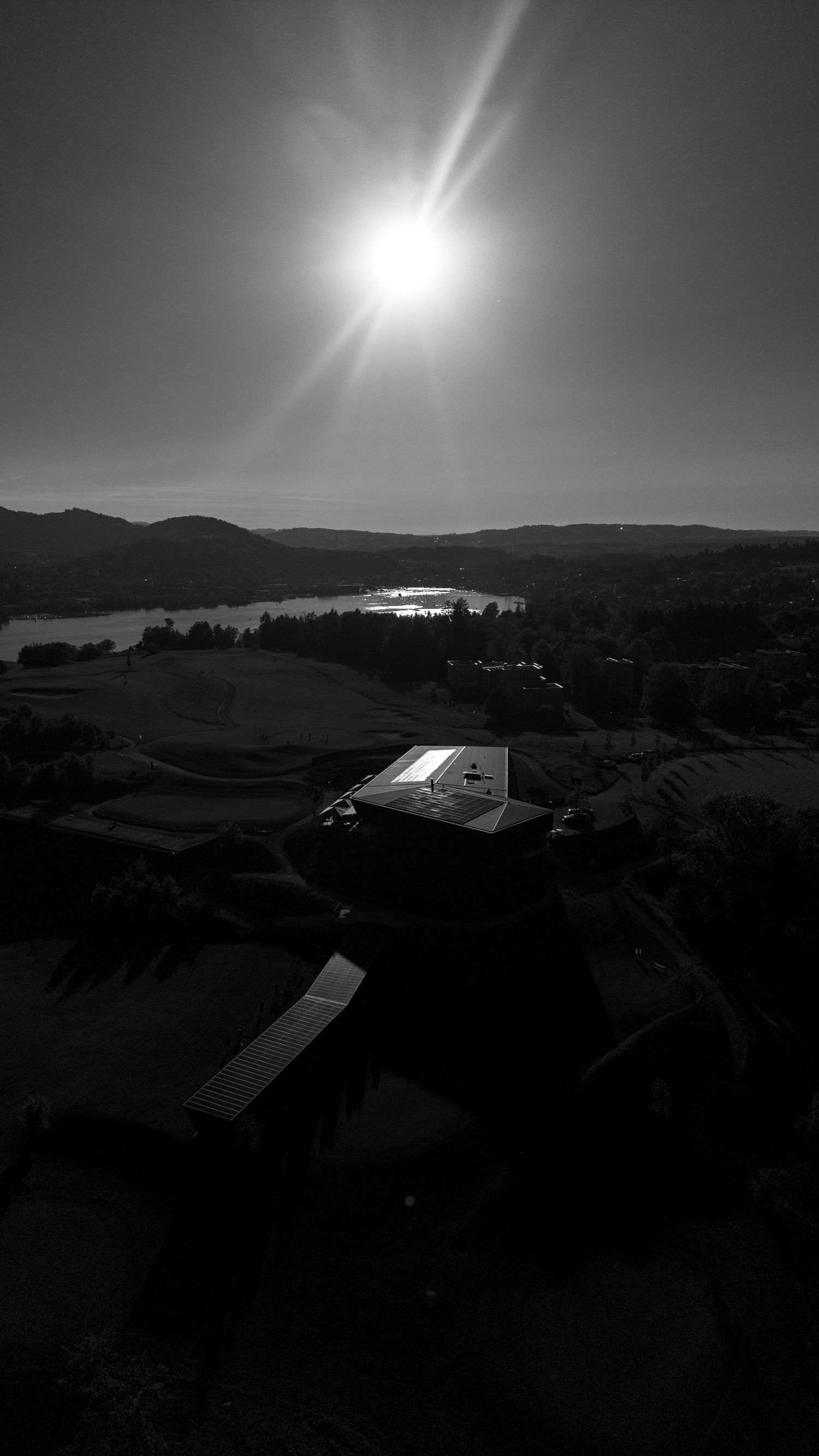 Aerial black and white photo of a building with solar panels, surrounded by trees and a lake, under a bright sunlit sky.