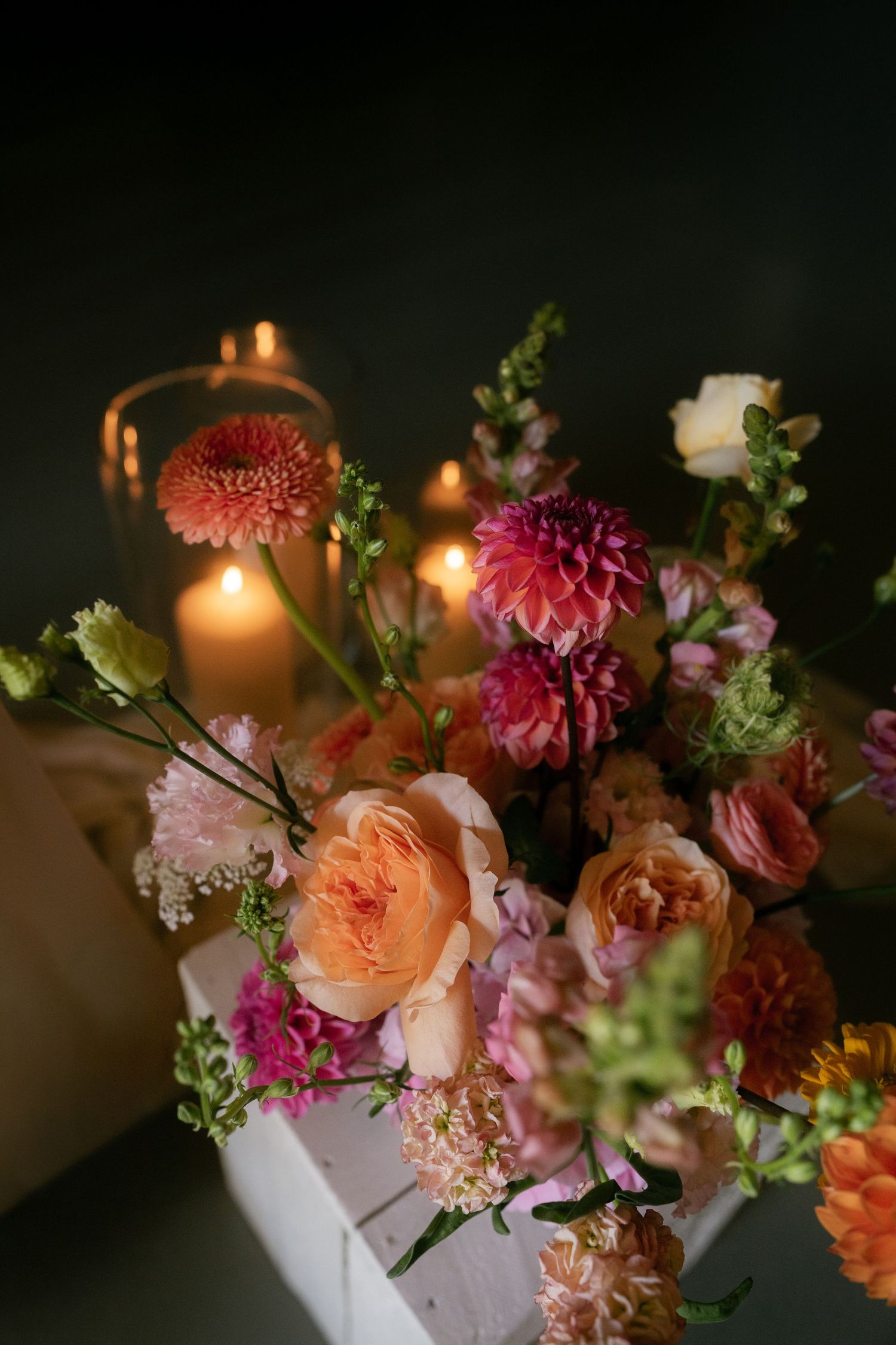Moody floral arrangement with dahlias, roses and greenery illuminated by candlelight.