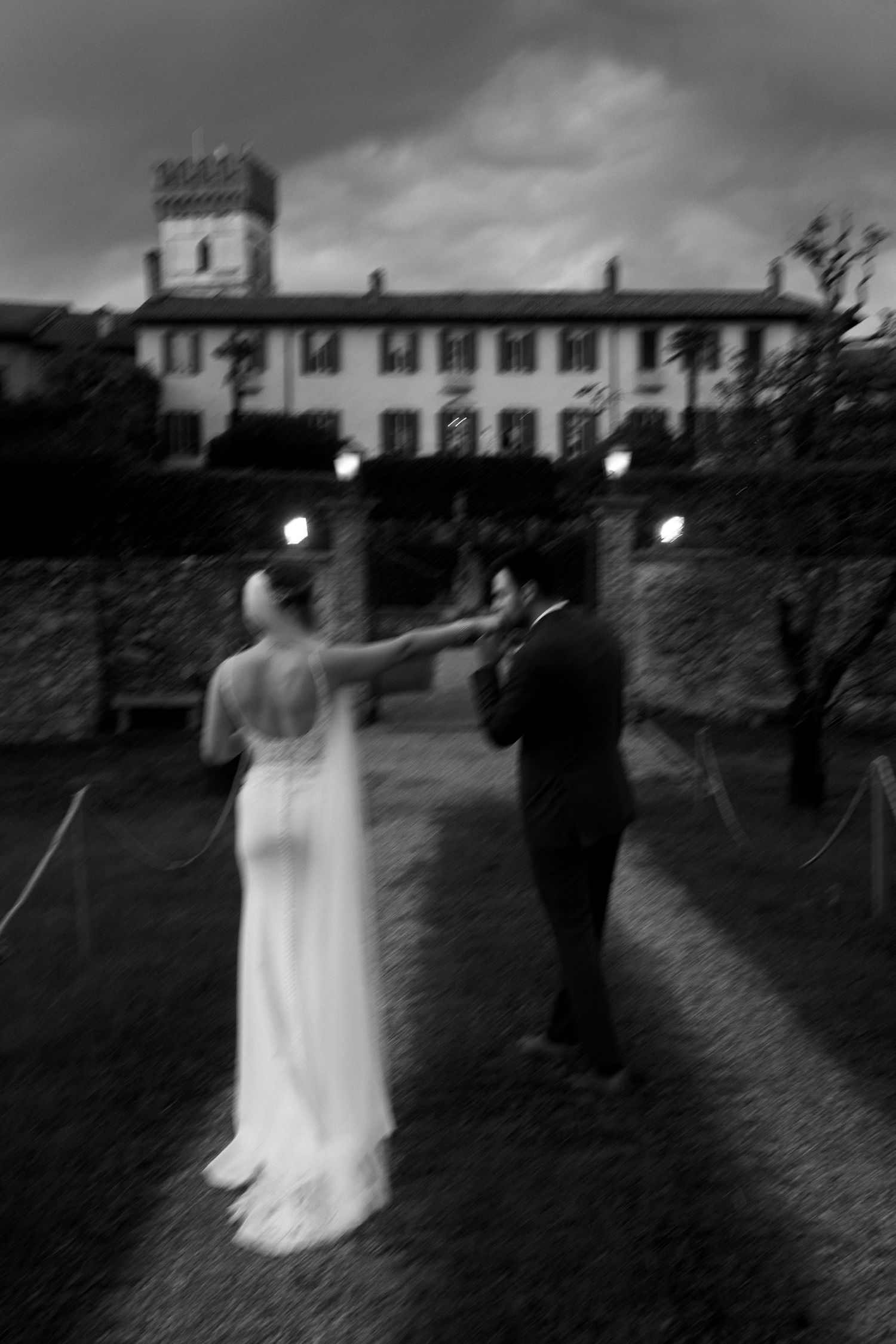 Blurred black and white photo of bride and groom dancing outside an elegant villa at dusk.