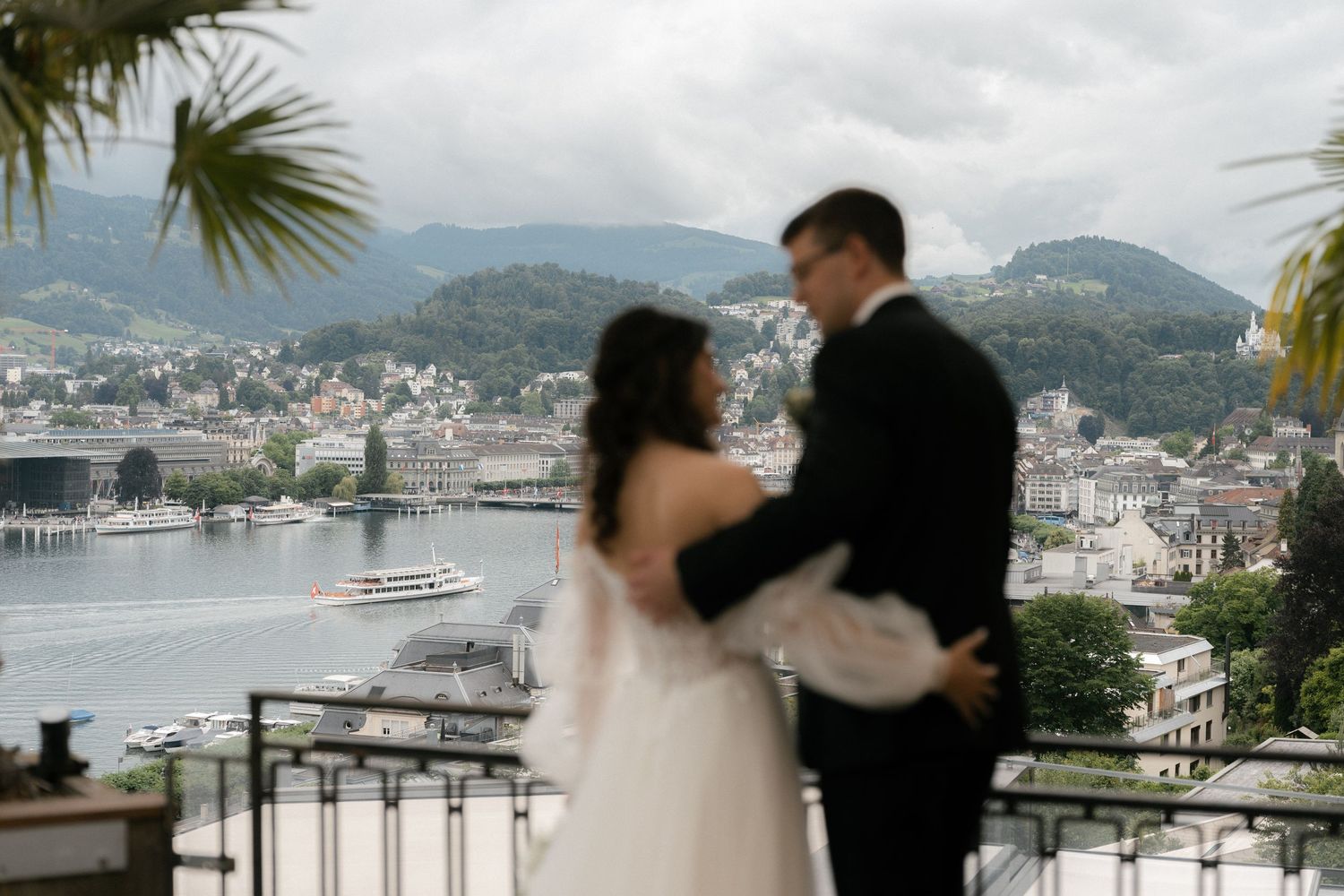 Bride and groom embrace on a balcony overlooking Lake Como with mountains and cityscape in background. Montana Luzern