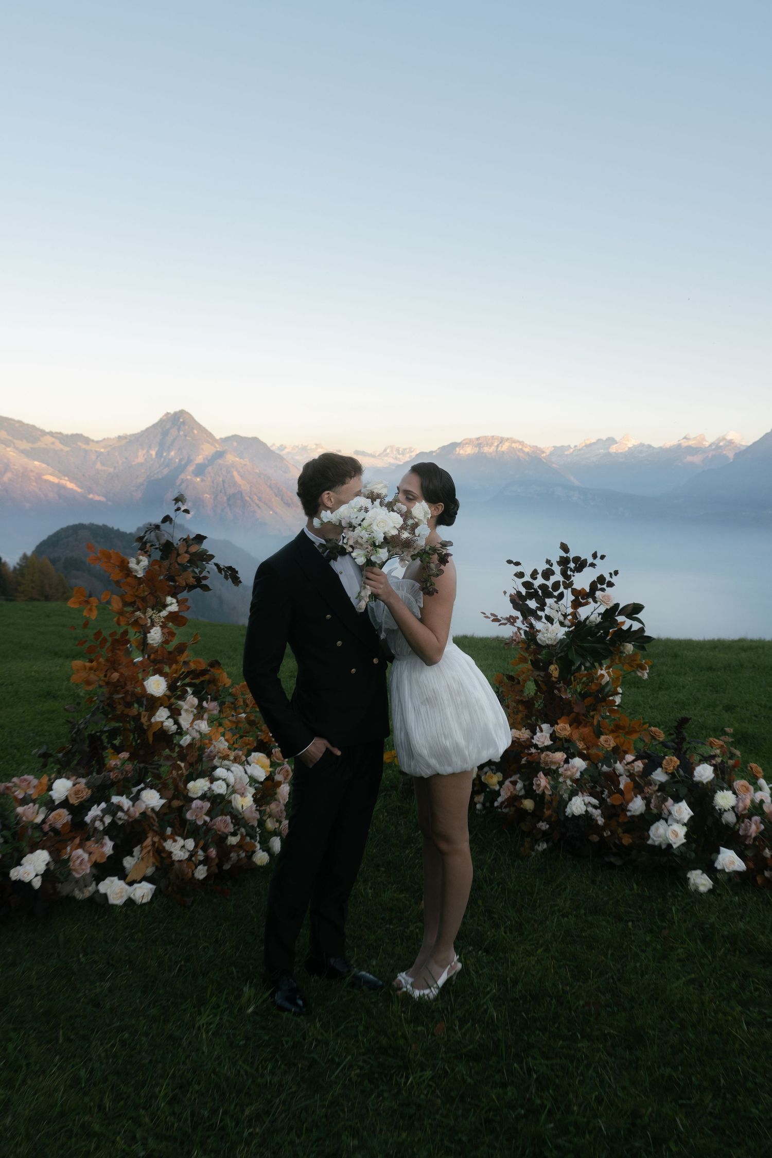 Couple in wedding attire kissing behind white bouquet with floral arch and mountain lake backdrop at dusk at Villa Honegg Bürgenstock