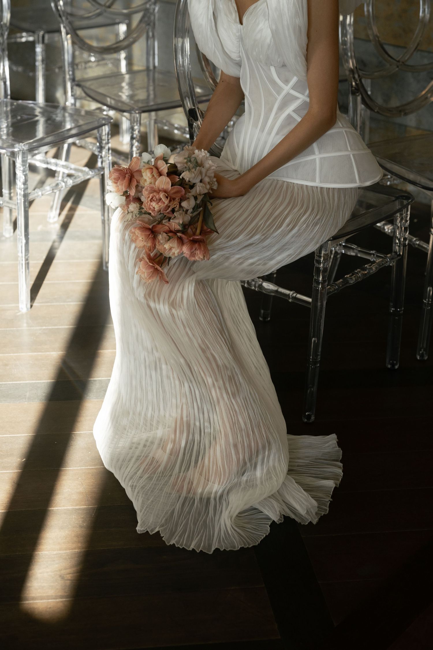 Bride in elegant white pleated wedding gown holding a bouquet of dried flowers in soft natural light.