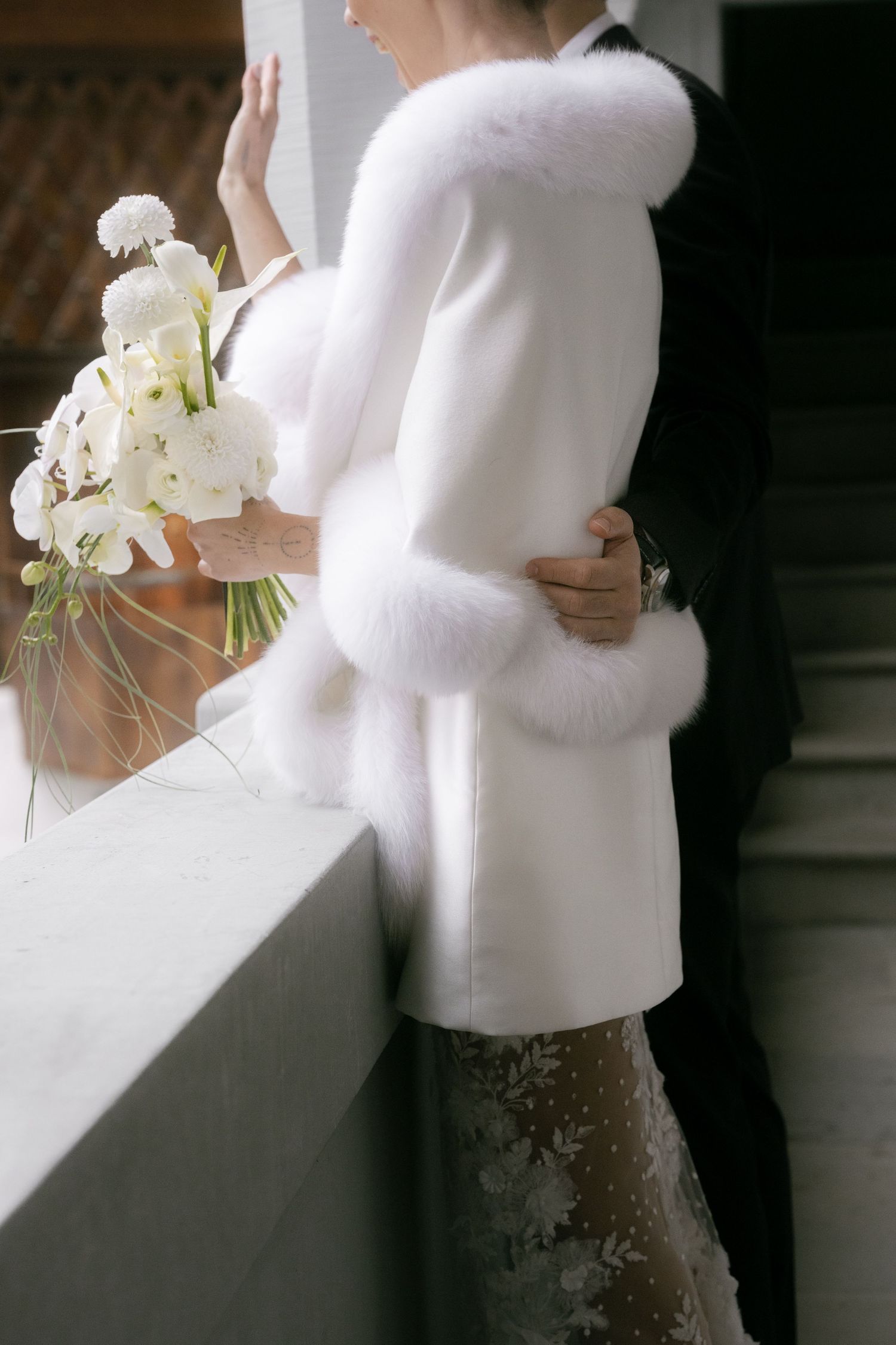 Bride in white fur-trimmed coat holding bouquet embraced by groom in black suit on elegant staircase.