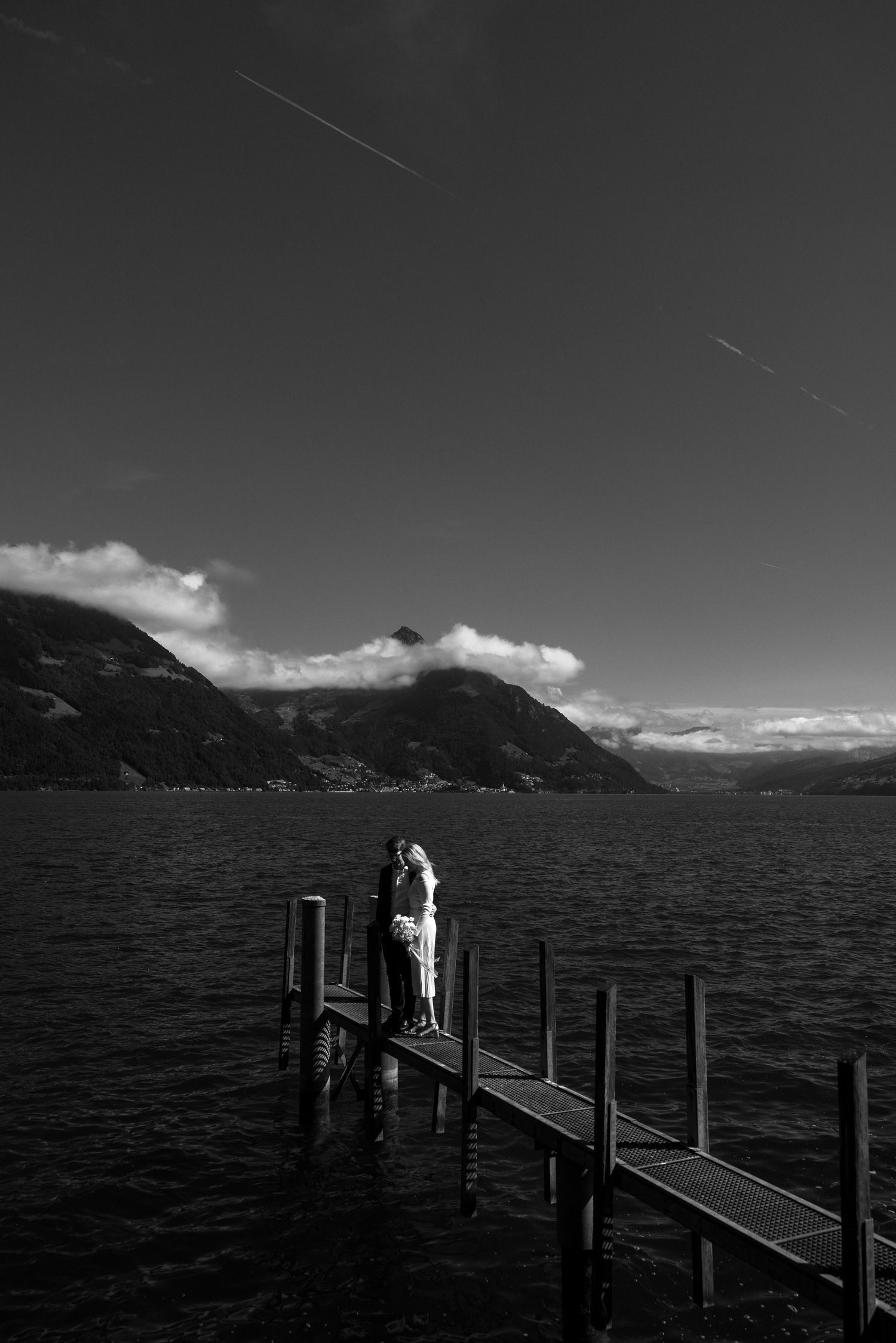 A black and white photograph shows a figure standing on a wooden pier overlooking a serene mountain lake.