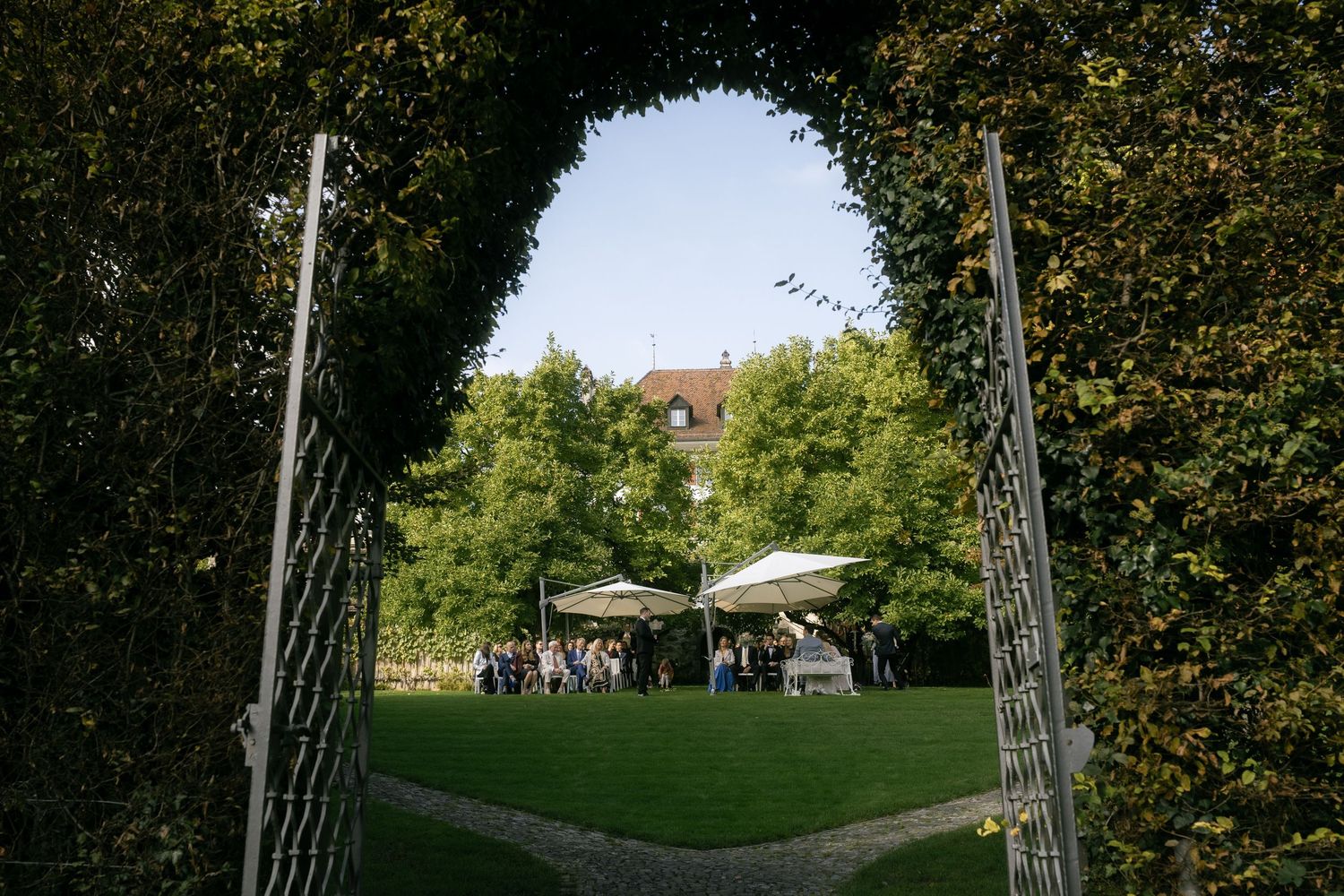 Garden party venue viewed through ornate iron gates with lush green trees and white umbrellas in background.