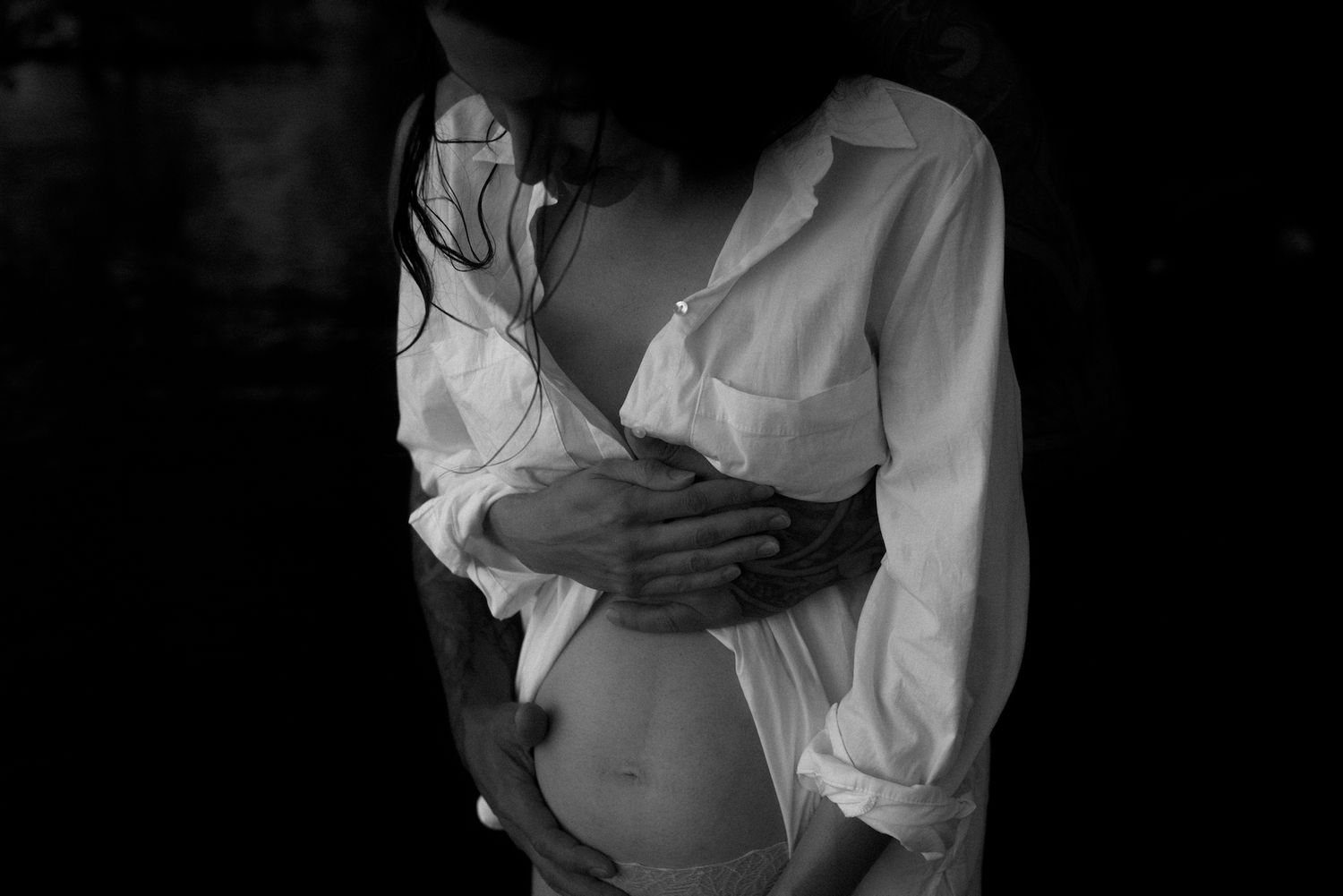 Intimate black and white maternity photo showing hands cradling a pregnant belly in a white button-down shirt.
