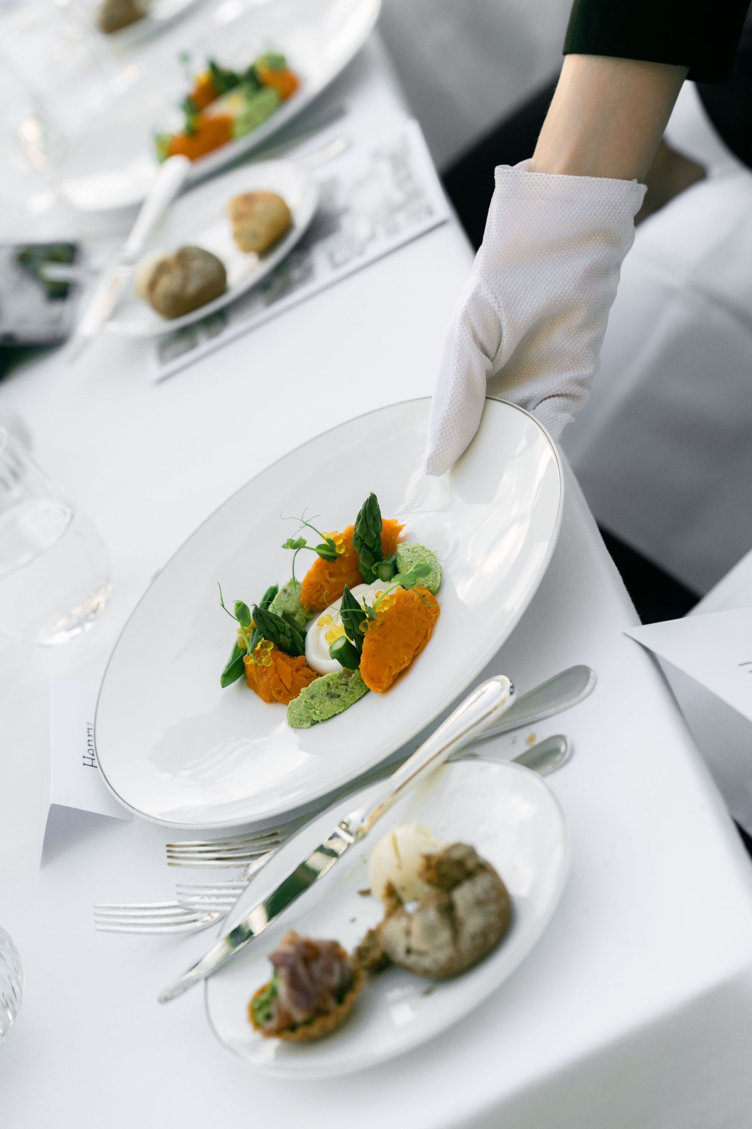 Gloved server placing elegant white oval plate with colorful gourmet appetizers on a formal white tablecloth setting.
