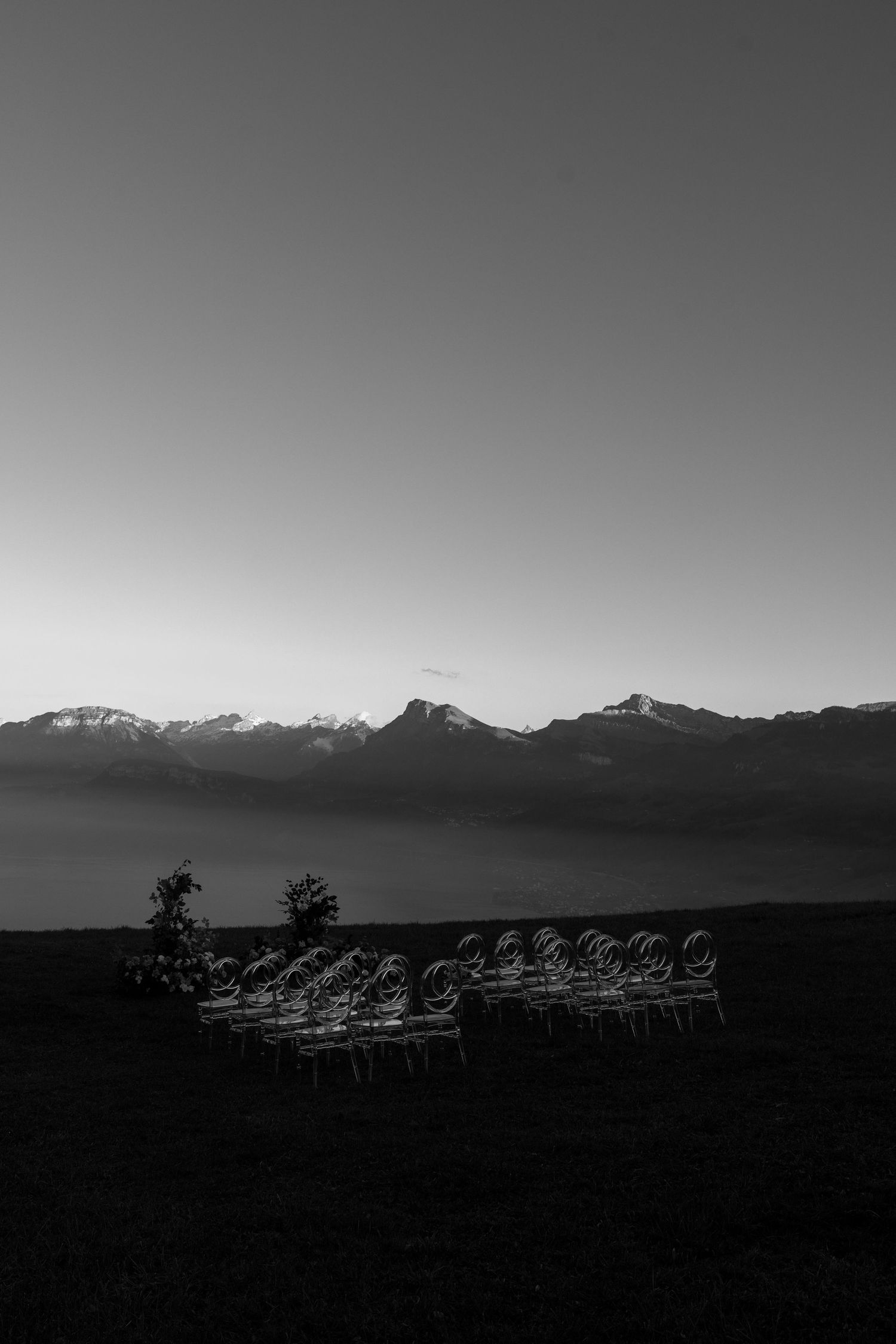 Black and white landscape photo of empty chairs arranged outdoors with snow-capped mountains in the background at dusk.