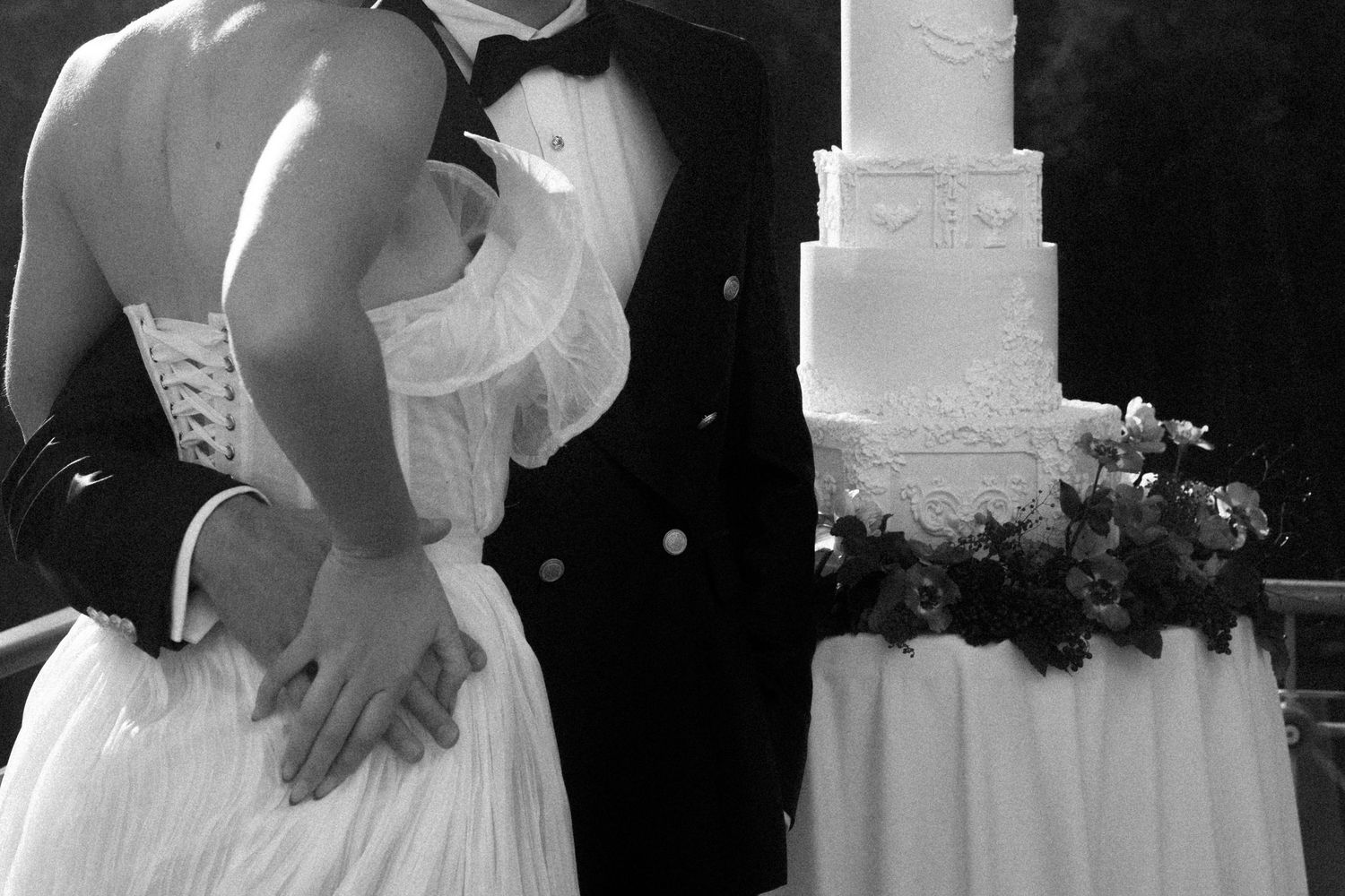 Black and white photo of a bride and groom embracing beside a elegant tiered white wedding cake with floral decorations.