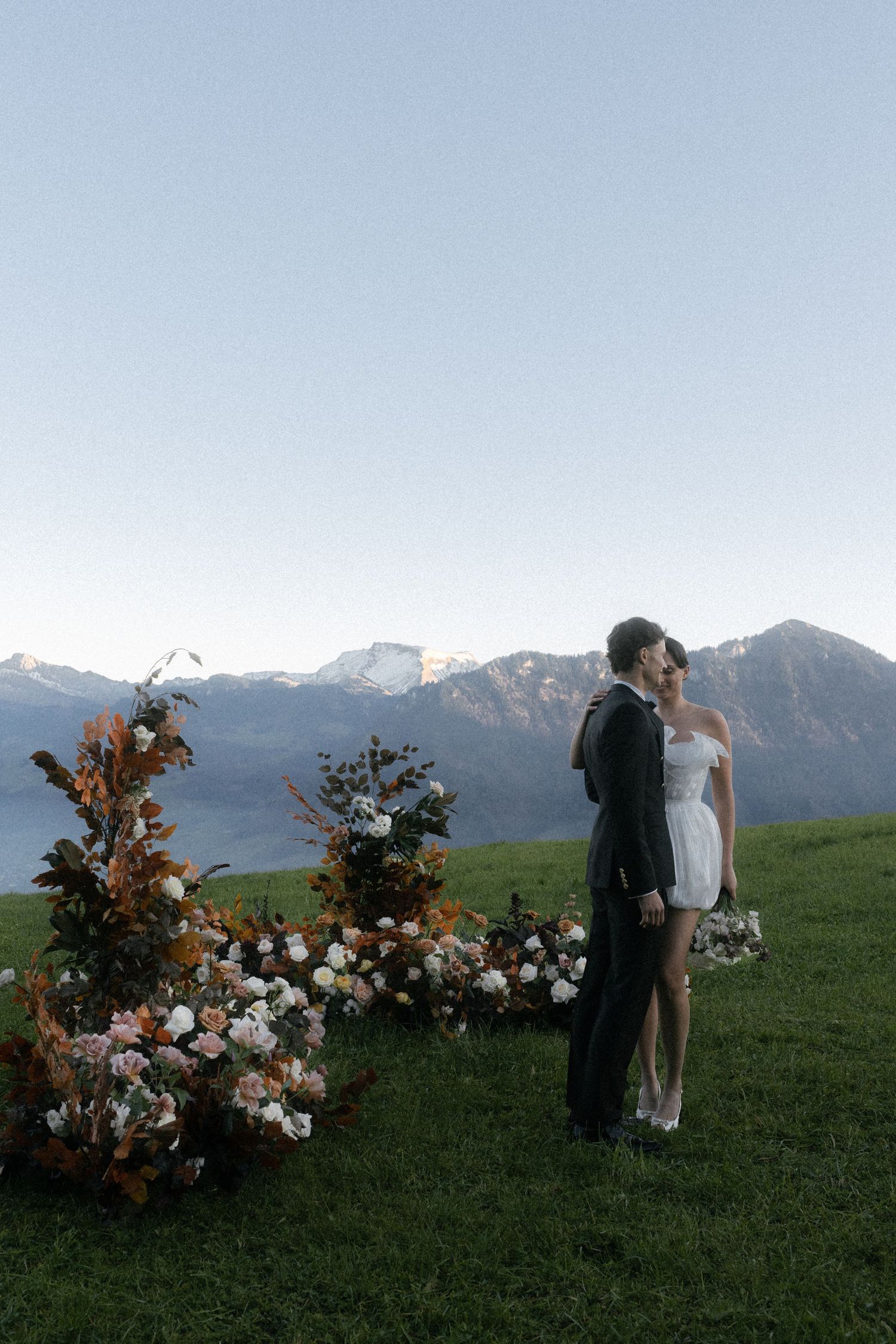Couple in wedding attire standing on green hillside with floral arrangements and snow-capped mountain backdrop at dusk. Villa Honegg Bürgens