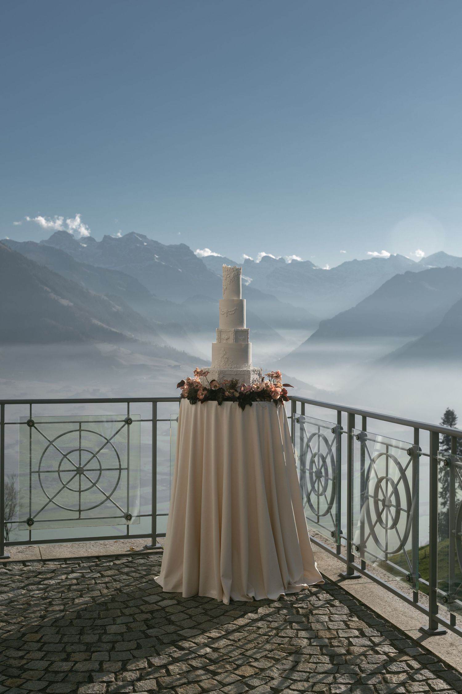 Elegant multi-tier wedding cake on draped table, balcony overlooking misty Alpine mountains and lake backdrop.