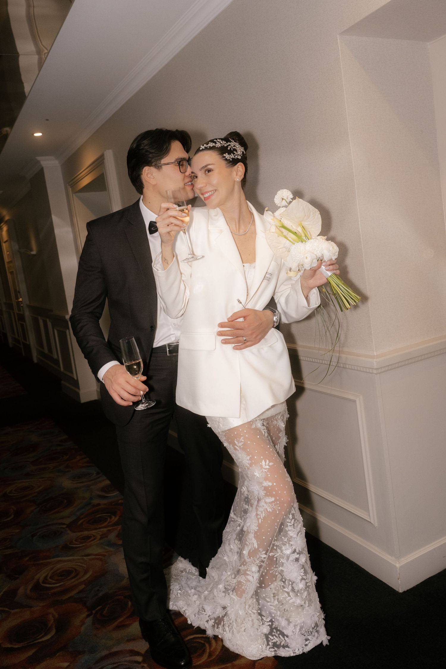 Elegant bride in white lace dress holding bouquet, groom in tuxedo holding champagne, sharing romantic moment in hotel corridor.