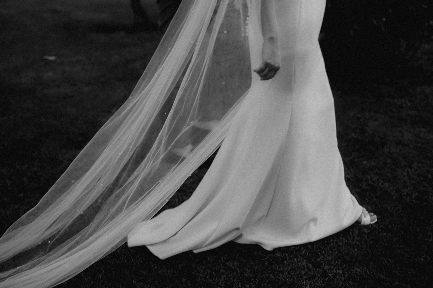 A flowing white wedding dress train and veil trail behind on a dark background, creating an ethereal bridal silhouette.