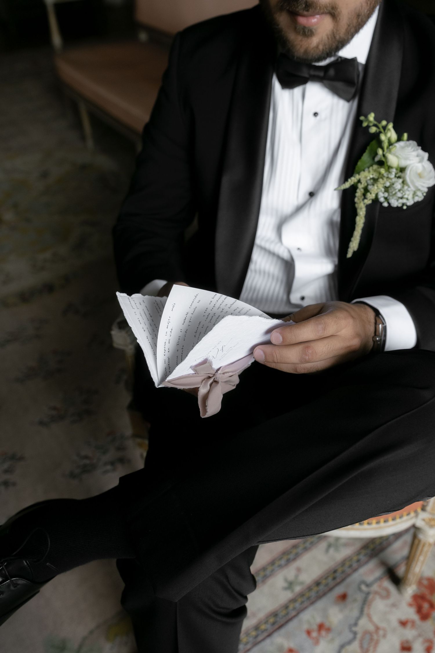 Groom in black tuxedo with floral boutonniere reads wedding vows from a ribbon-tied booklet while seated.