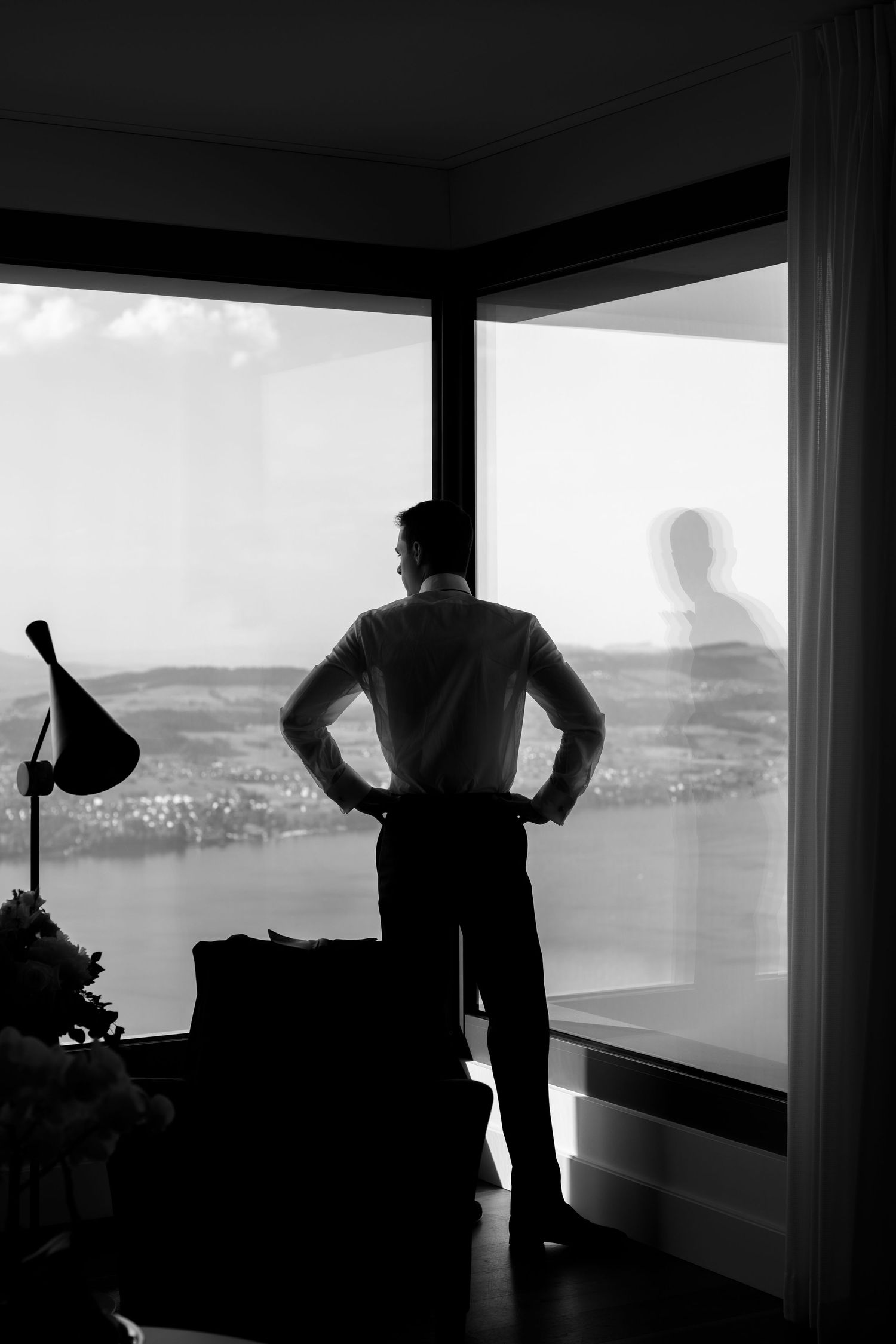 Black and white silhouette of a man standing with hands on hips before large windows overlooking a city.