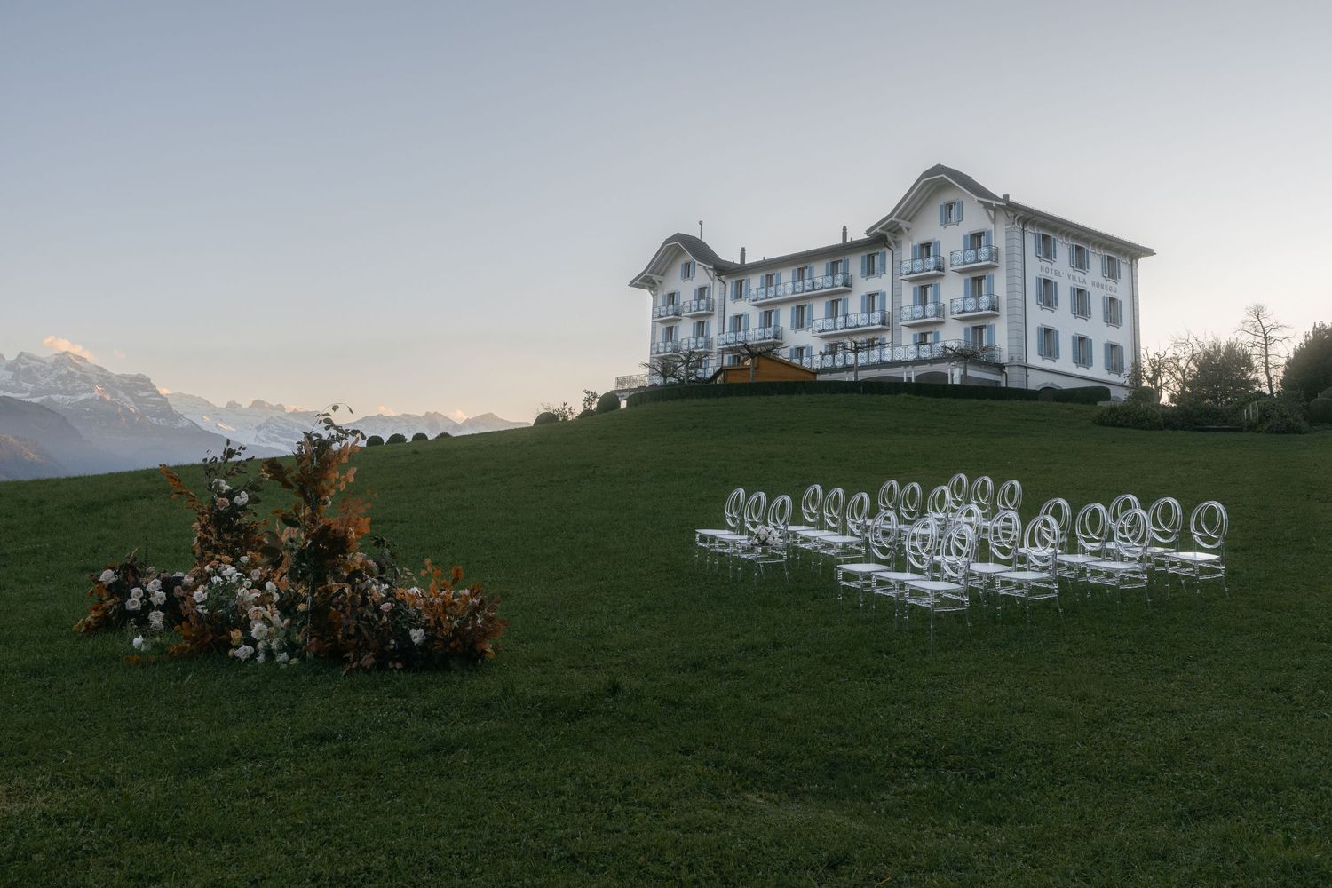 Outdoor wedding ceremony setup on a green hillside with floral arrangements and clear chairs, elegant white mansion Villa Honegg Bürgenstock