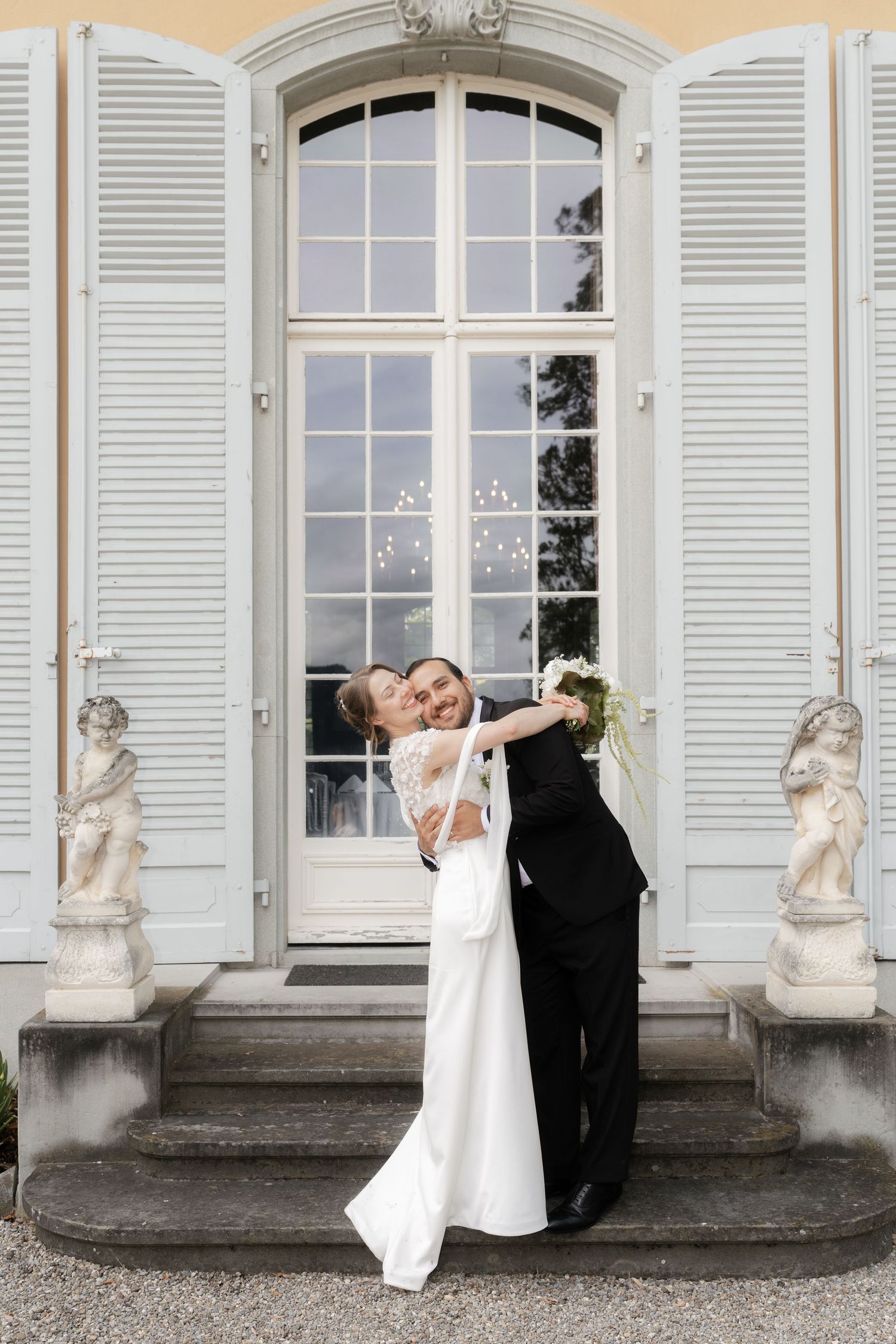 Bride and groom embracing joyfully on steps of elegant villa with white shutters and cherub statues.