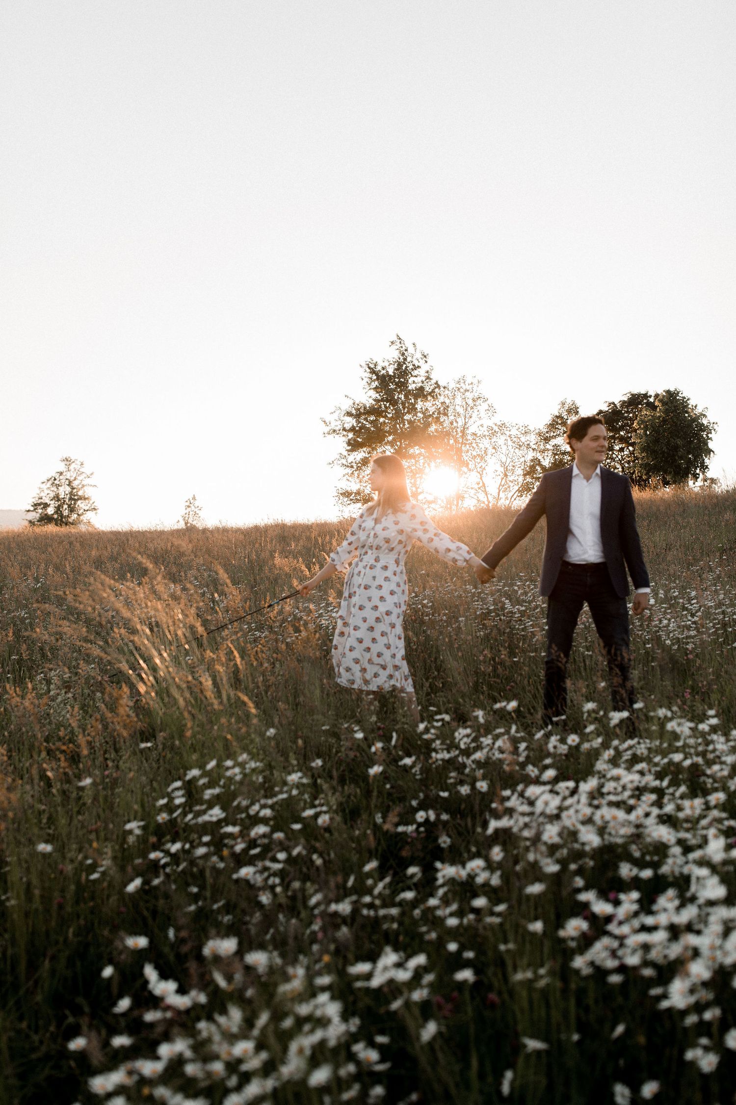 A couple holds hands in a wildflower meadow at sunset with white daisies blooming in the foreground.