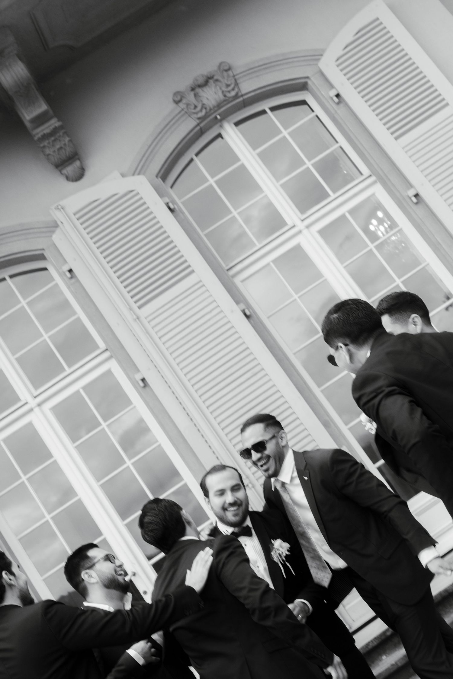 Black and white photo of groomsmen in suits laughing and celebrating together outside elegant building with shuttered windows.