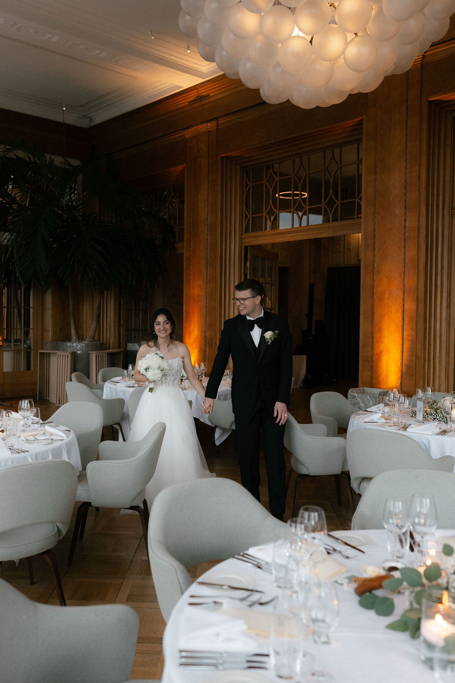 Bride and groom walk hand in hand through an elegantly decorated wedding reception venue with white balloon chandelier. Montana Luzern