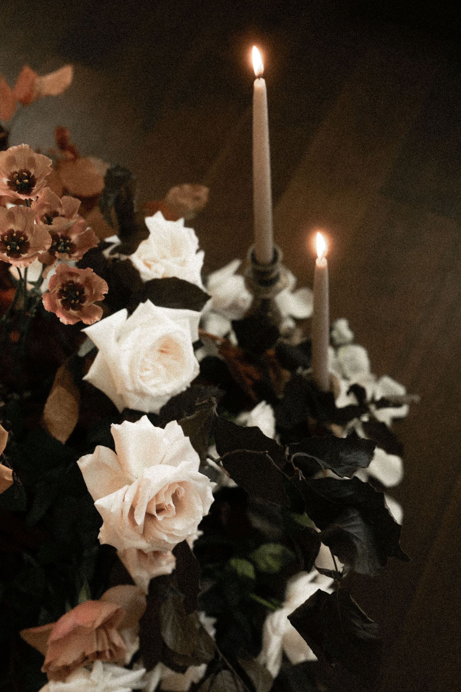 Elegant floral arrangement with white and blush roses, dark foliage, and two lit taper candles in a moody, romantic setting.