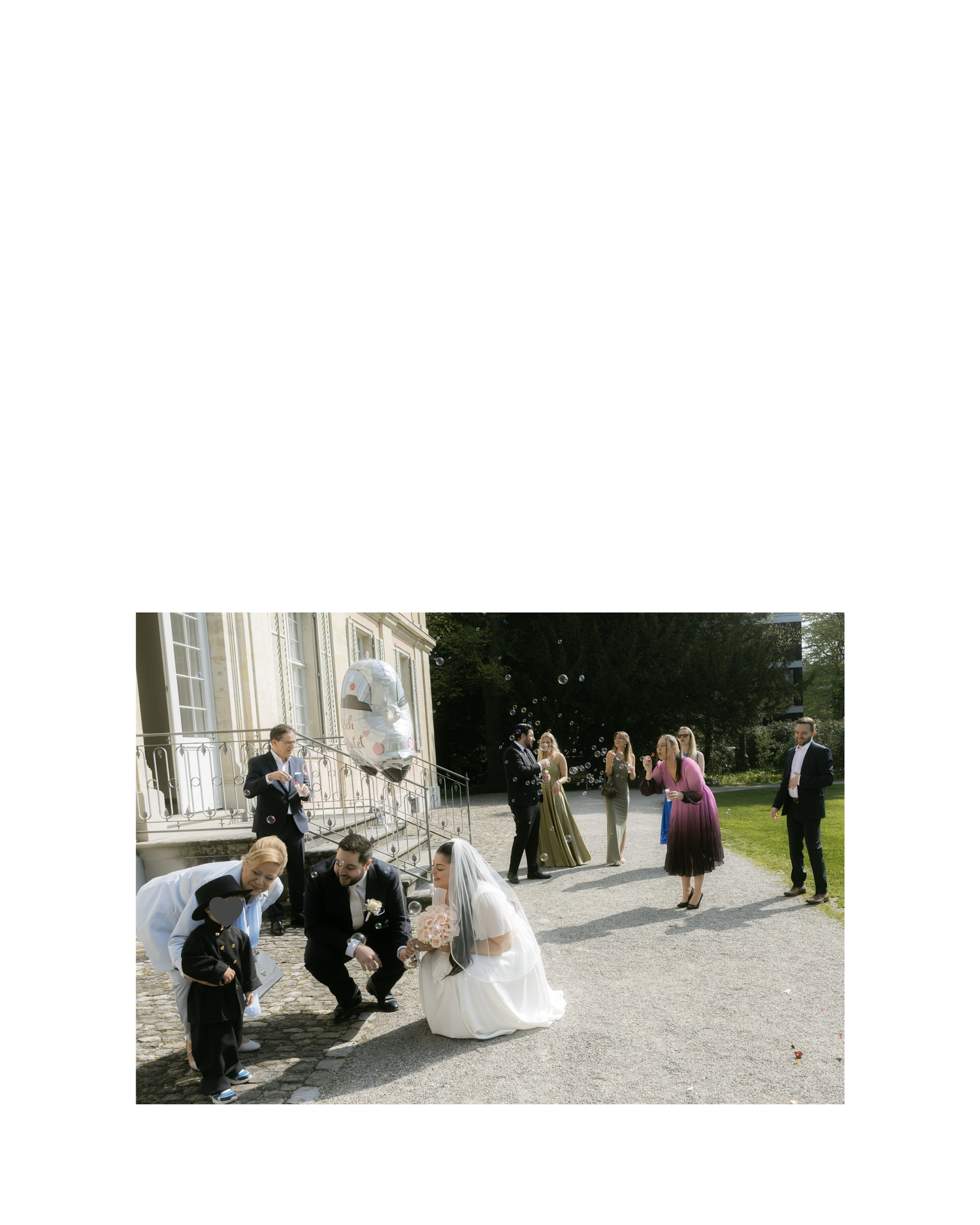 Wedding party poses outdoors near elegant building, bride in white gown crouches with guests while others stand nearby.