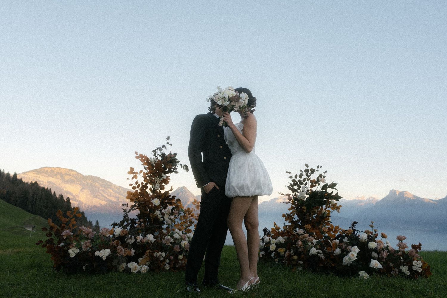 Couple kissing at outdoor mountain wedding ceremony with floral arrangements and Alpine peaks at dusk. Villa Honegg Bürgenstock