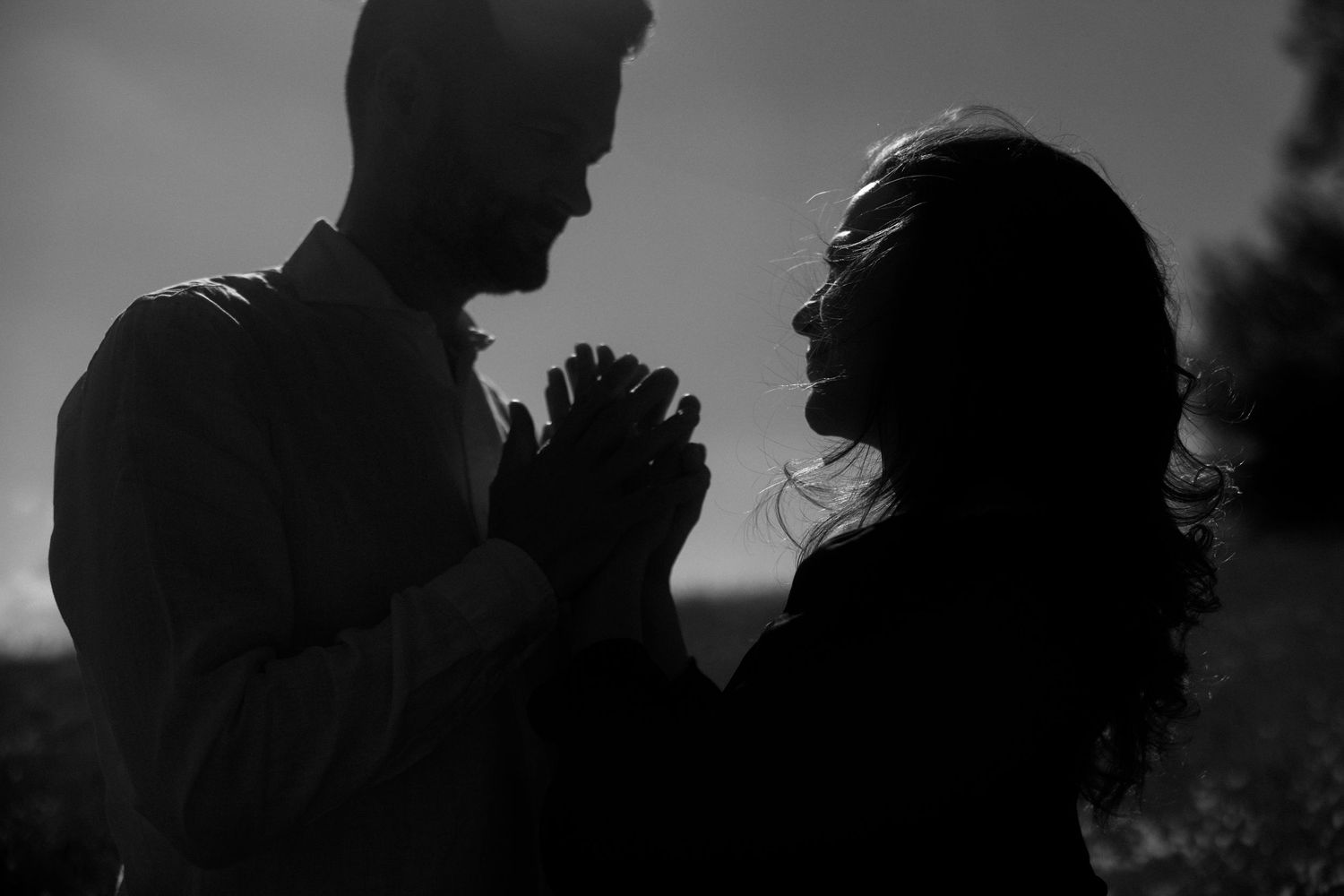 A silhouetted couple shares an intimate moment in black and white as they face each other with clasped hands.
