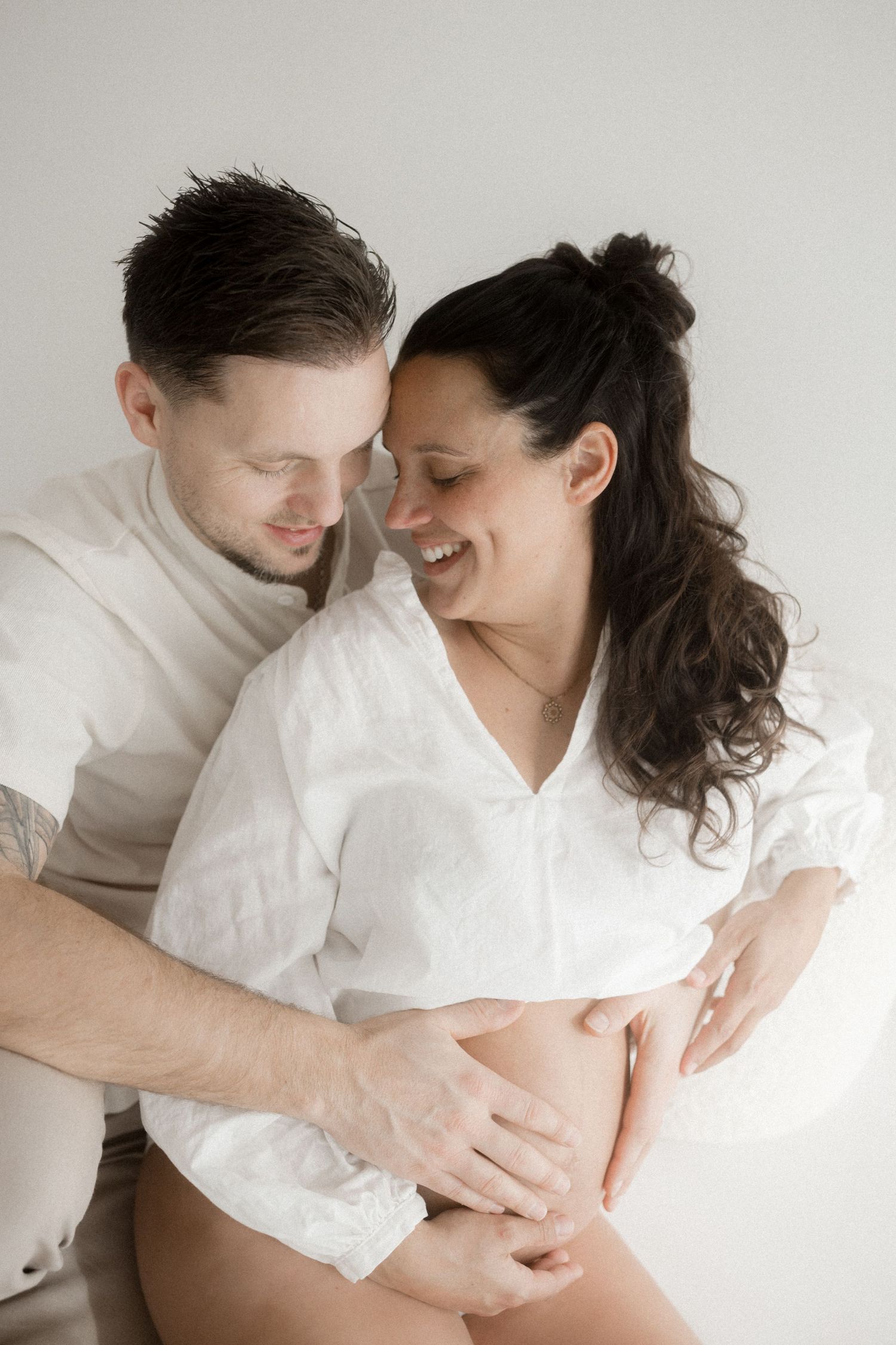 Intimate maternity photoshoot series with couple in white clothing against light background.