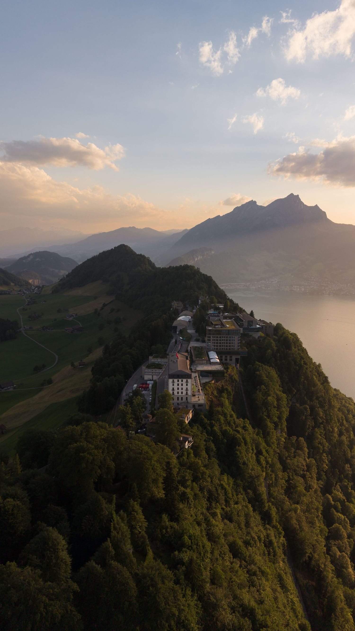 Aerial drone view of a hilltop village surrounded by lush green forests overlooking a serene alpine lake at sunset.
