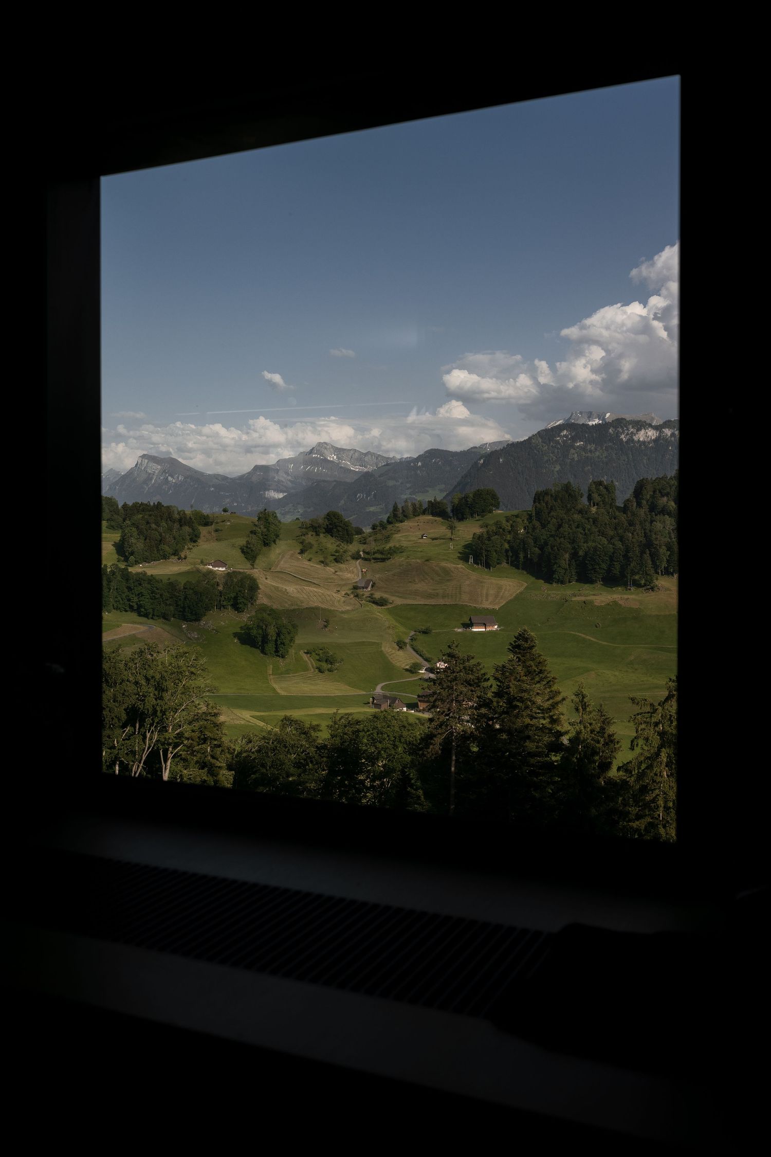 View through a dark window frame of lush green Alpine meadows, scattered farmhouses, and snow-capped mountains under blue sky.