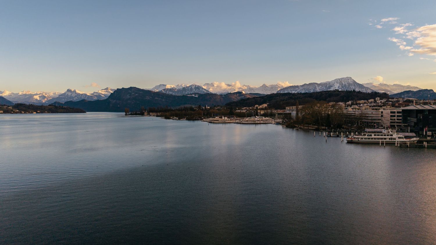 Panoramic view of snow-capped mountains reflecting in a calm lake with boats docked along the shoreline at sunset.