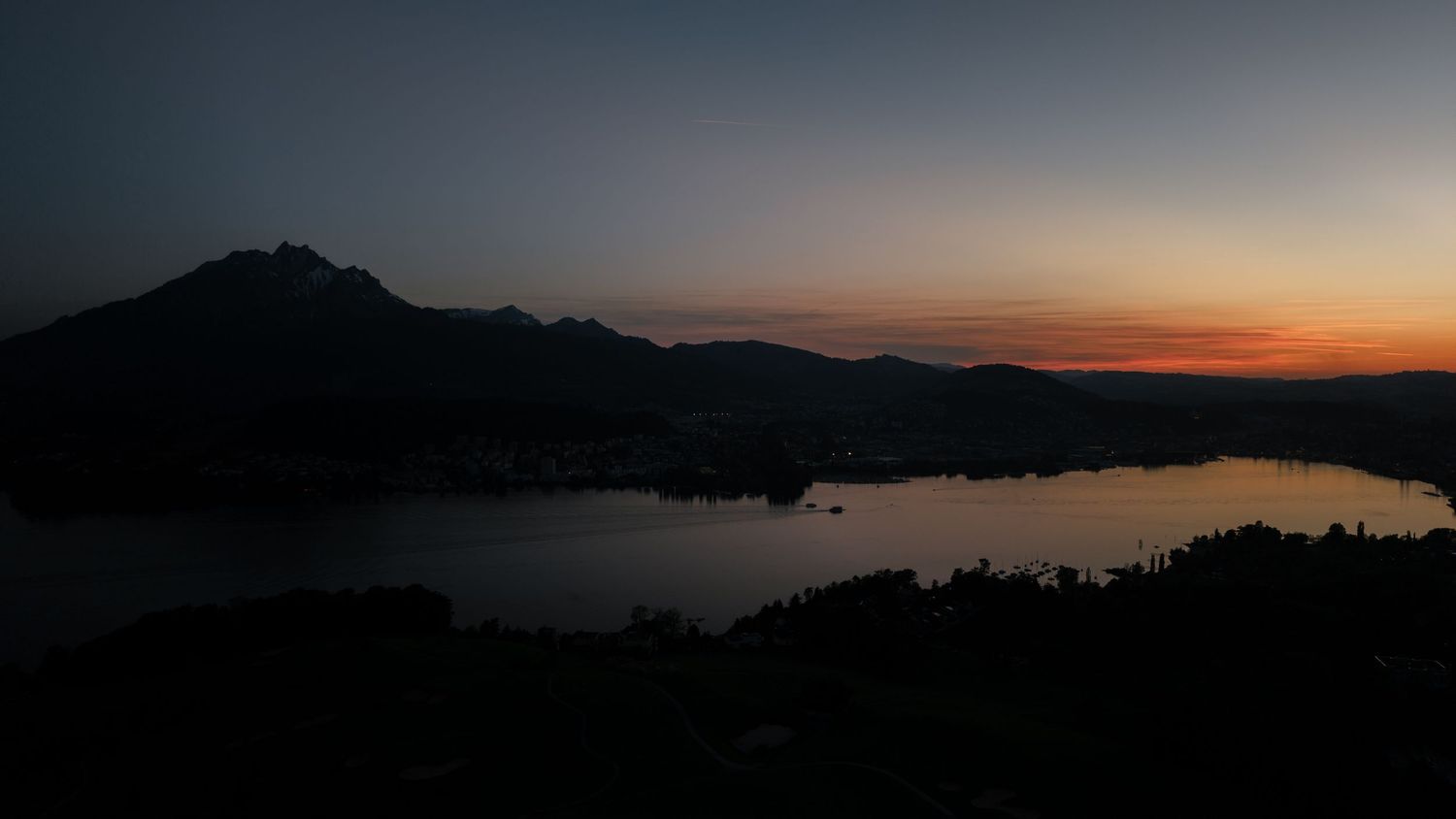 Dramatic sunset silhouette over a tranquil mountain lake with warm orange horizon glow reflecting on calm water.